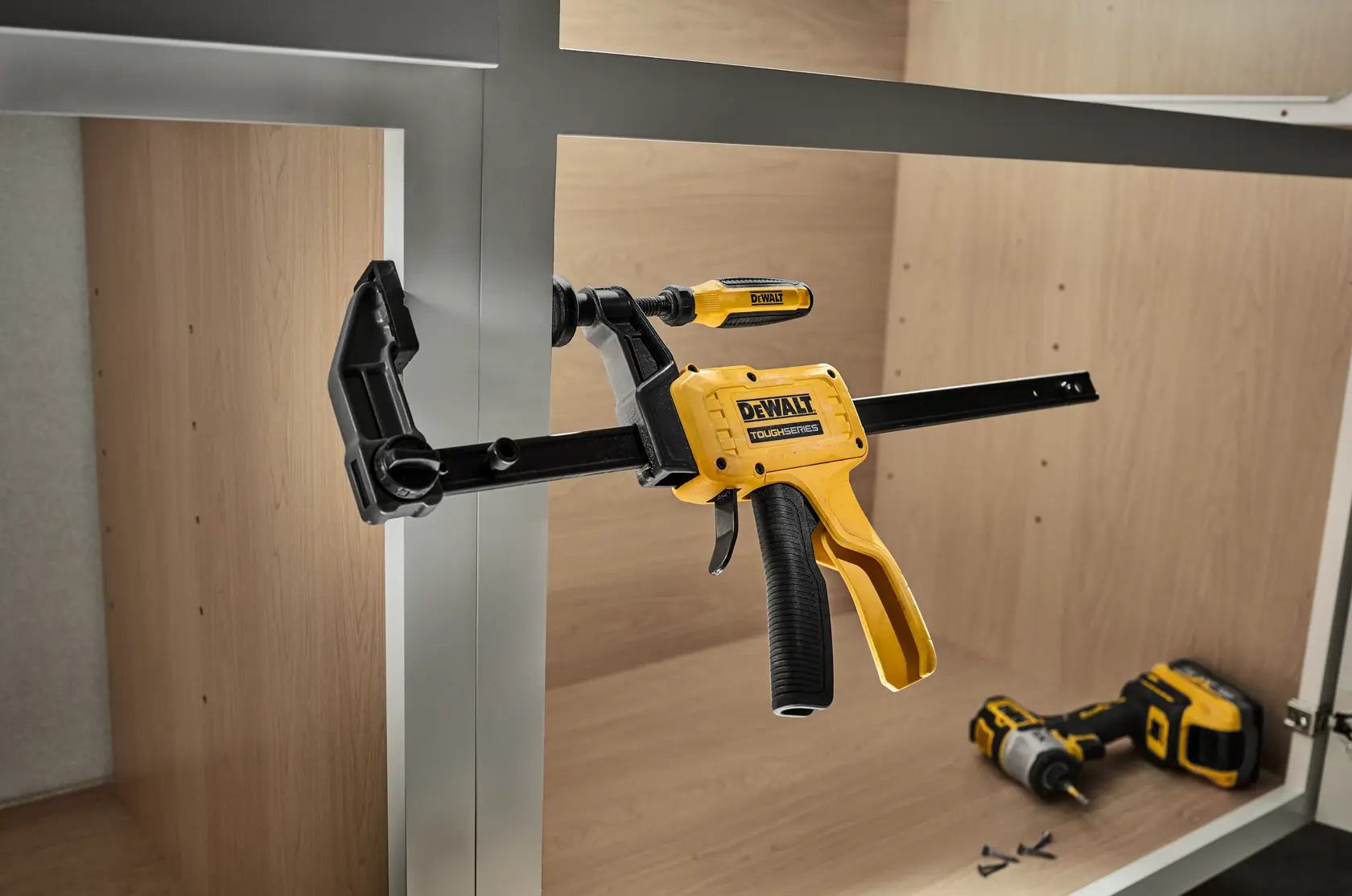 A DEWALT ToughSeries trigger clamp is being used to hold two metal frames together inside a wooden cabinet. A DEWALT cordless power tool and some screws are visible on the cabinet floor in the background.