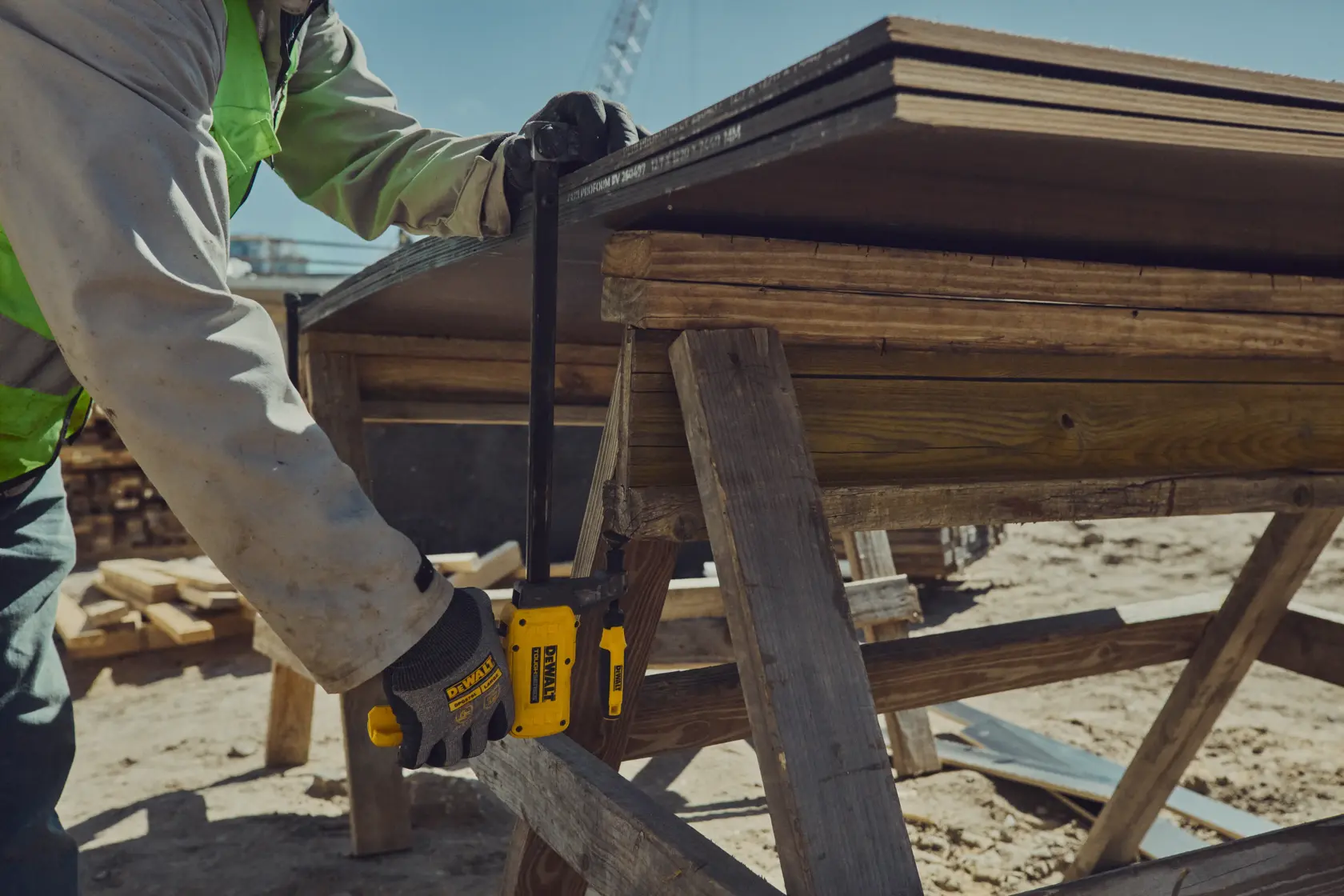 A close-up of a person wearing gloves and a safety vest using a DEWALT DWHT83600 clamp to secure wooden boards at a construction site.