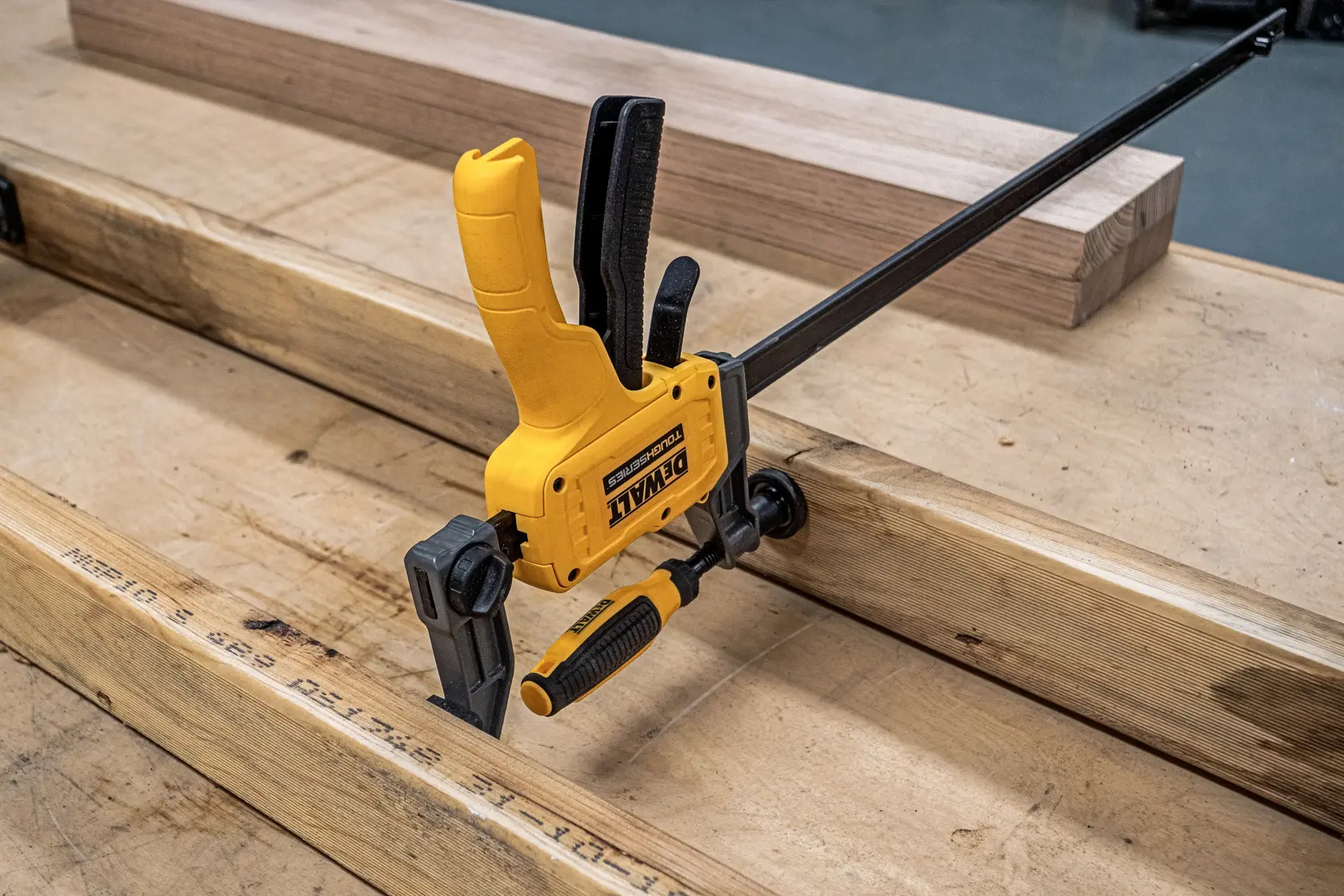 A yellow and black DEWALT quick-release bar clamp is holding two pieces of wood together on a workbench in a woodworking setting.