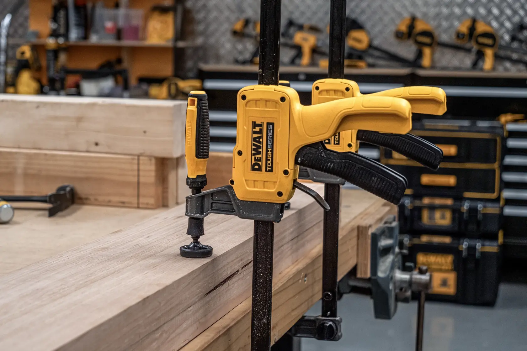 A DEWALT ToughSeries quick-change bar clamp is securing a piece of wood to a workbench in a workshop. The clamp is yellow and black and is being used in a woodworking setting with several other tools and equipment visible in the background.