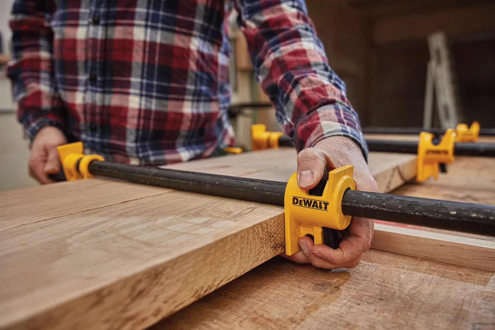 A person wearing a plaid shirt uses a DEWALT pipe clamp to secure wooden boards together on a workbench.