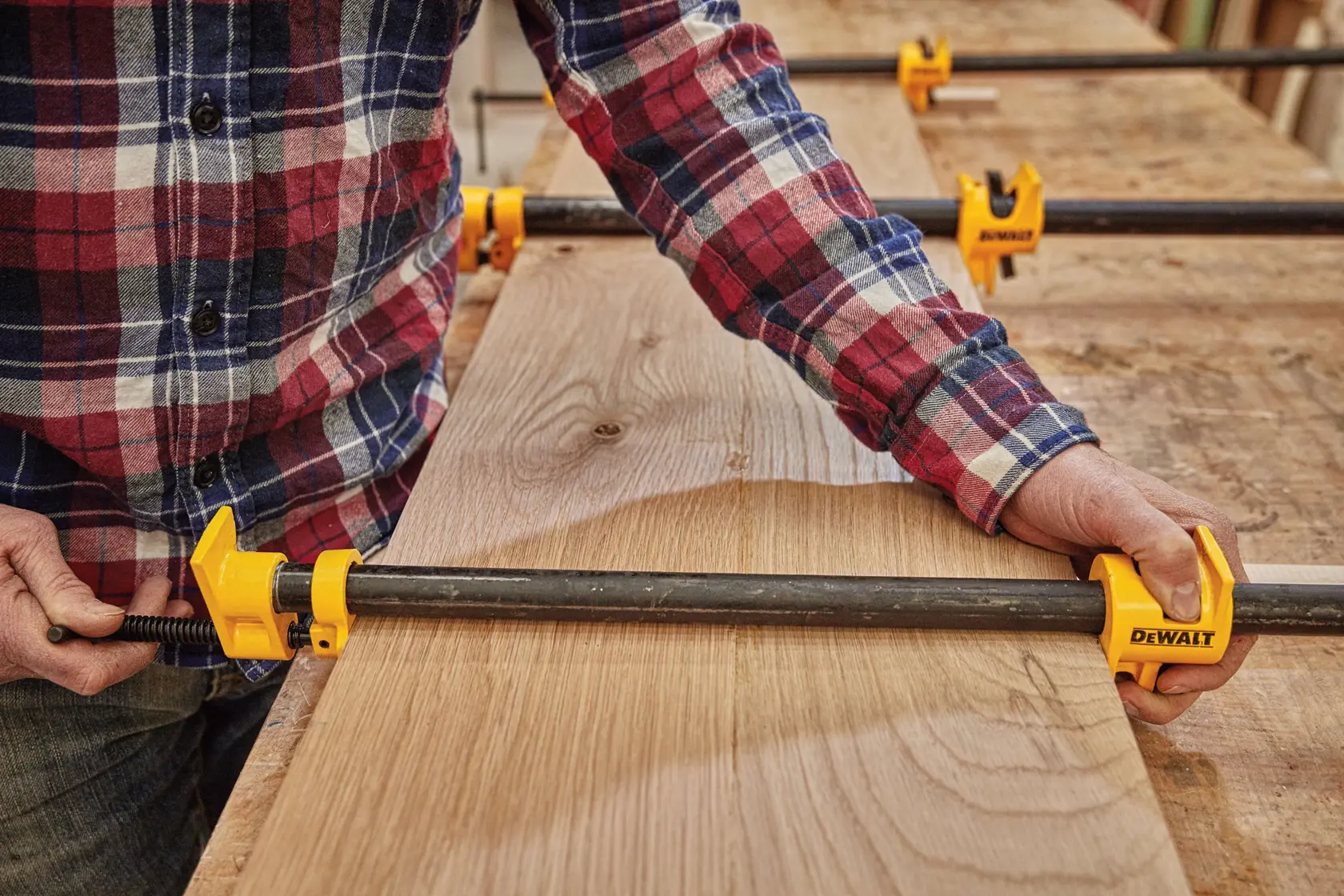 A person wearing a plaid shirt uses a DEWALT pipe clamp to secure wooden boards on a workbench. Several DEWALT clamps are visible holding the wood together.