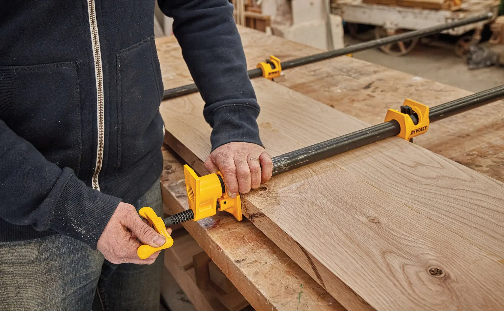 A person using yellow DEWALT pipe clamps to secure wooden boards on a workbench in a workshop.