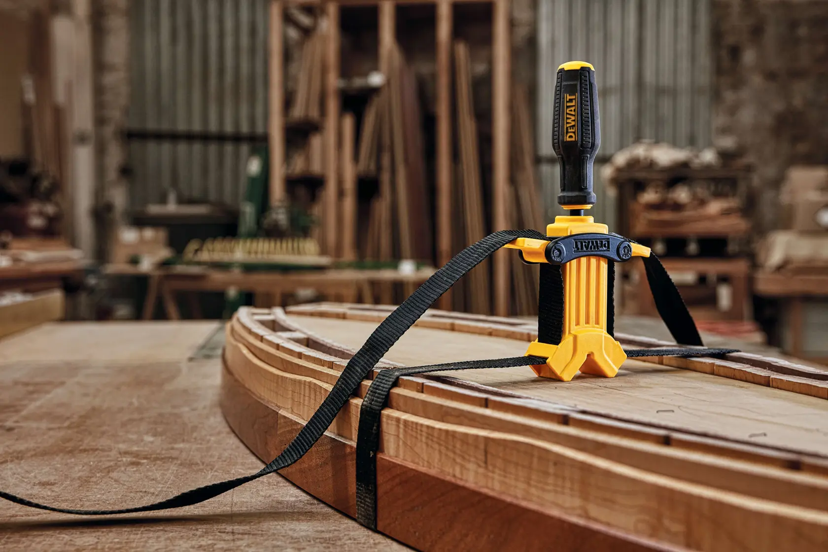 A DEWALT strap clamp is securing several curved wooden pieces together on a workbench in a woodworking shop, with a black strap tightening the assembly.