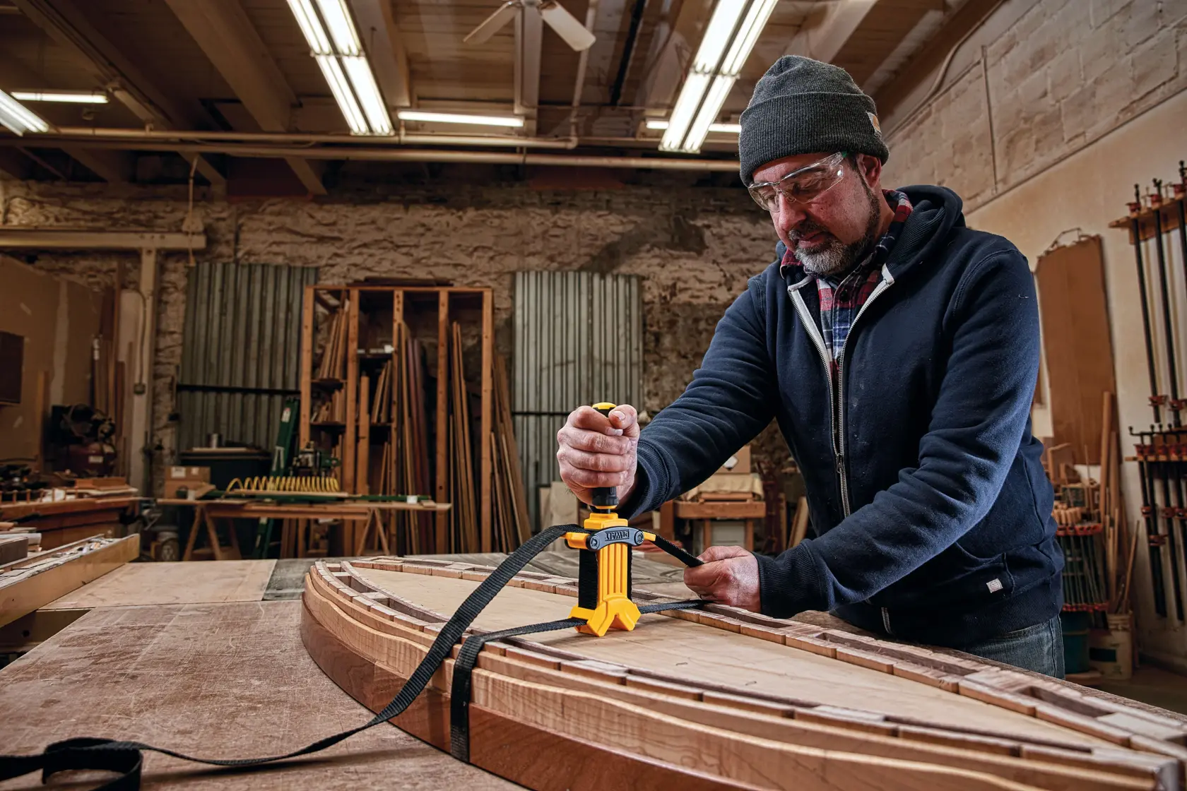 A person using a DEWALT strap clamp (model DWHT83839) to secure wooden boards together on a workbench in a woodworking shop.