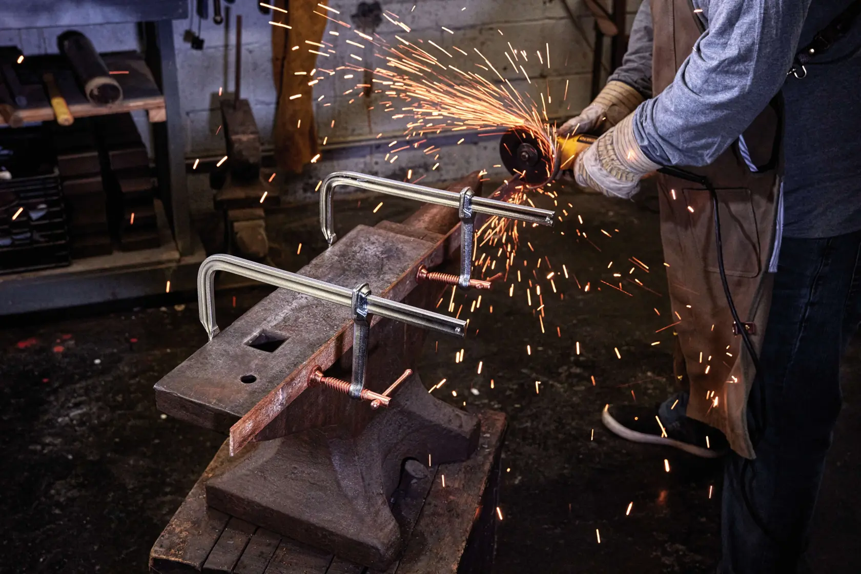A person using a power tool to cut metal, with sparks flying. The metal is clamped securely to an anvil with two DEWALT C-clamps. The person is wearing protective gloves and apron, and the workspace has tools and equipment in the background.