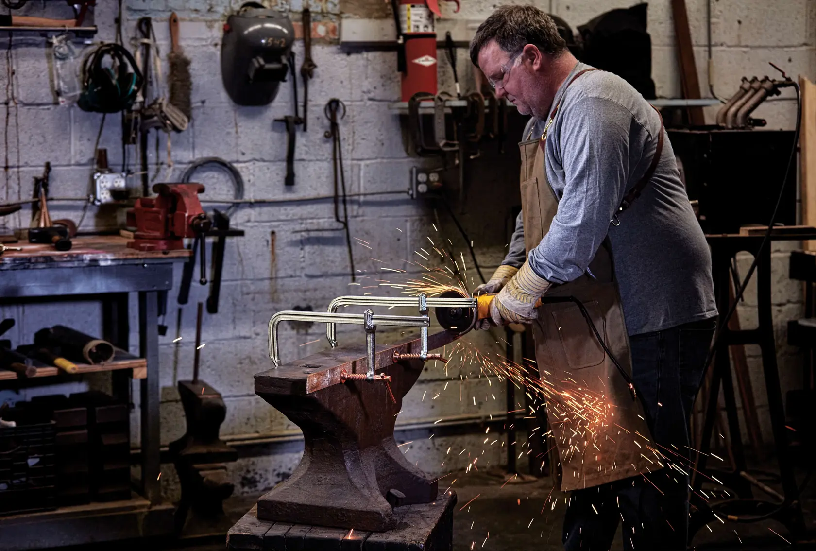 A person in a workshop is using a DEWALT C-clamp (SKU DWHT83850) to hold a metal piece on an anvil while grinding it with a power tool, creating sparks. The person is wearing protective gloves and an apron.