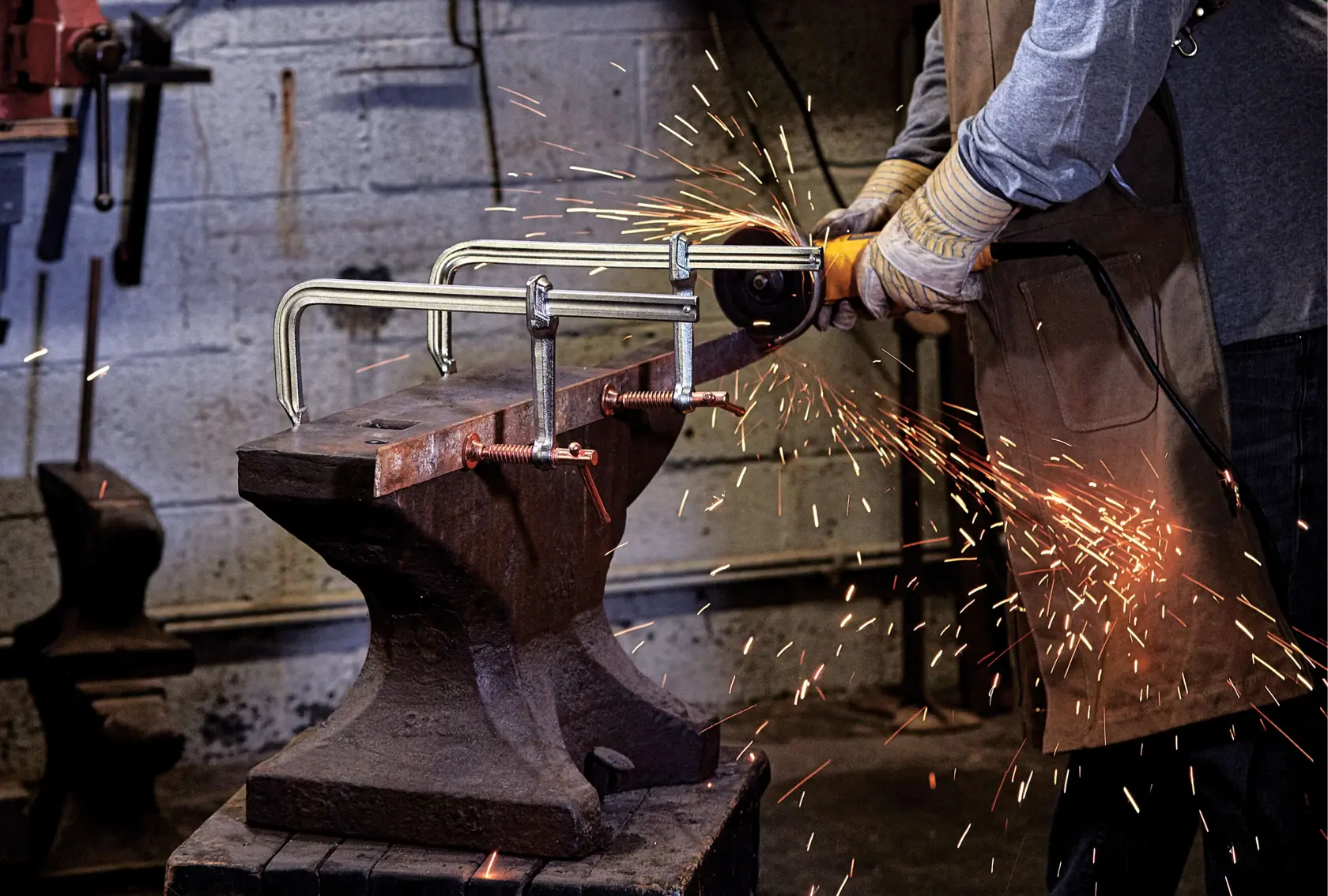 A person uses a DEWALT power tool to grind a metal object secured with clamps to an anvil, producing sparks in a workshop setting.