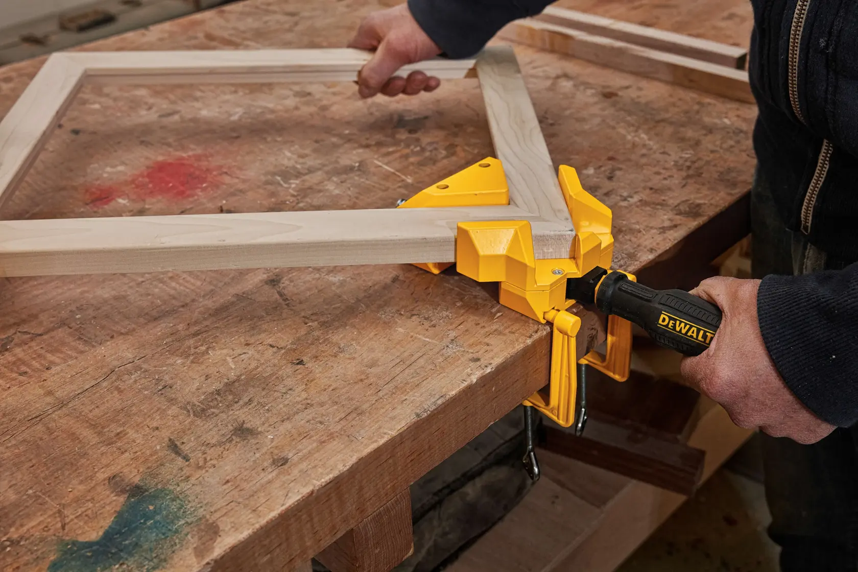 A person uses a DEWALT corner clamp to hold two wooden pieces together on a workbench, assisting in assembling a wooden frame.