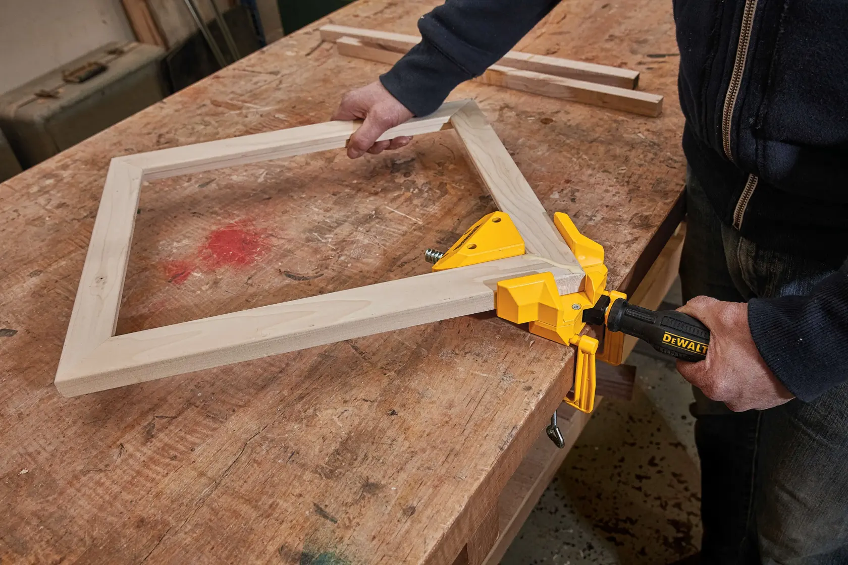 A person uses a DEWALT corner clamp on a wooden frame while working on a woodworking project on a workbench.