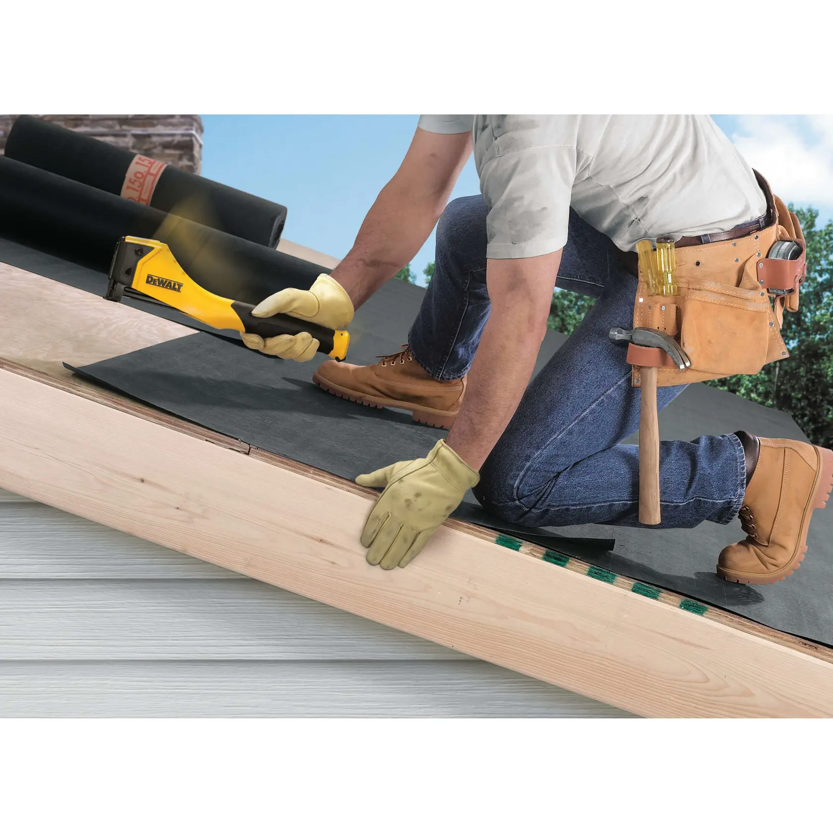 A person wearing work gloves, boots, and a tool belt is kneeling on a roof, using a yellow and black DEWALT staple gun to attach roofing material. Rolls of roofing material are visible in the background.