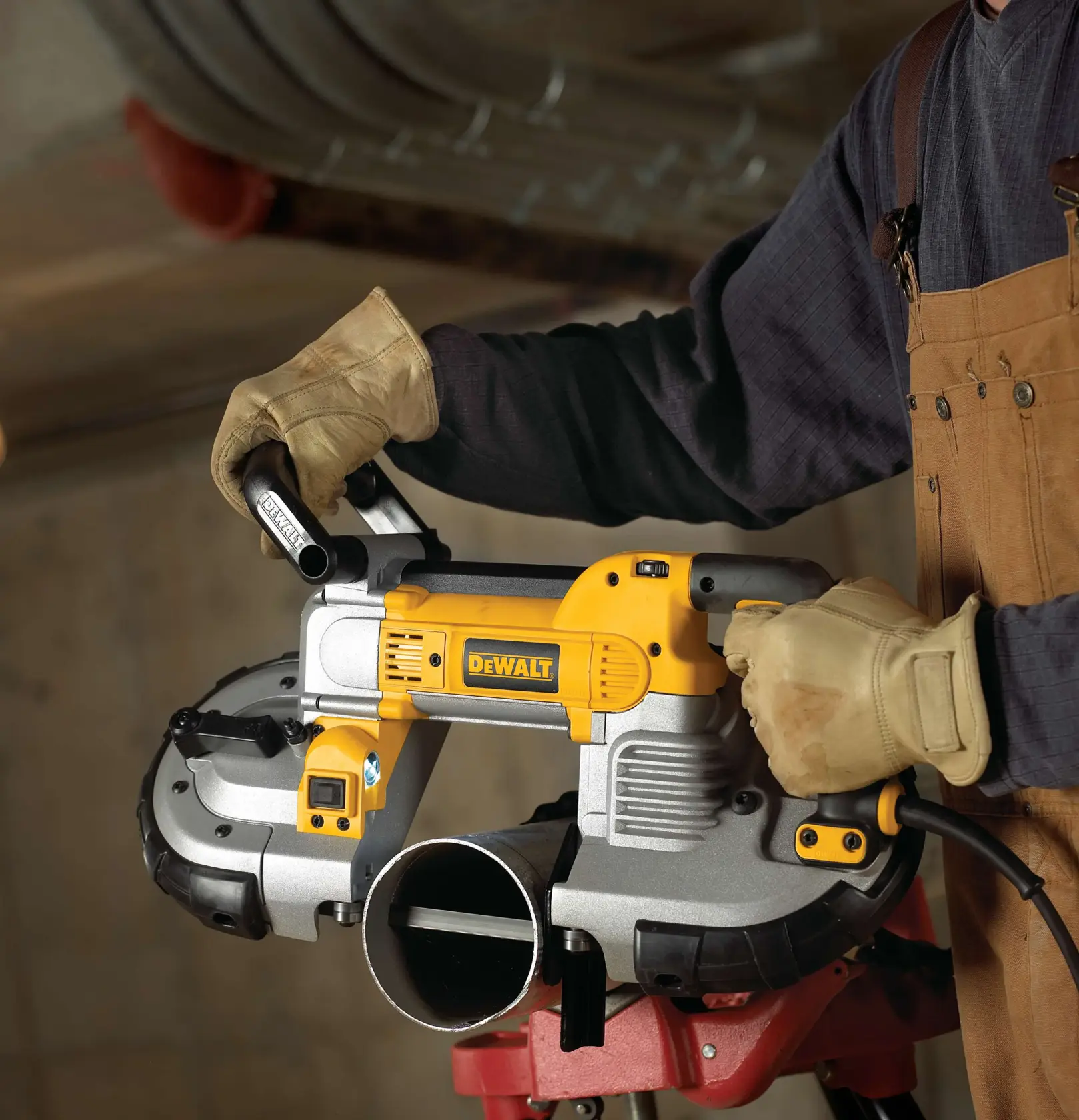 A person wearing gloves and work overalls is using a DEWALT DWM120 portable band saw to cut through a metal pipe in an industrial setting.