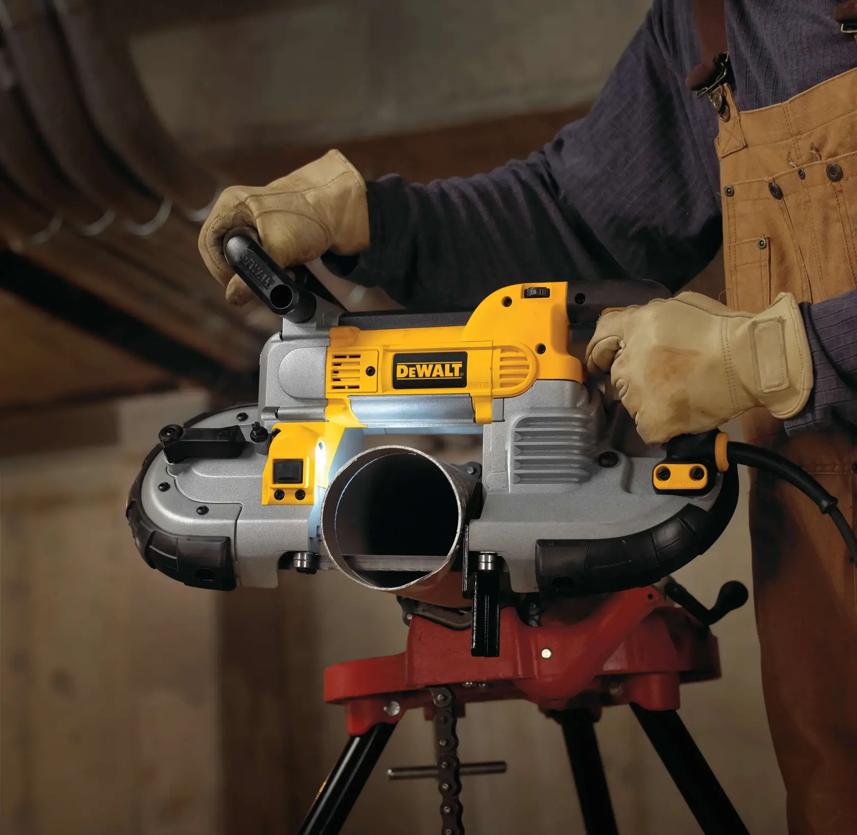 A person wearing work gloves uses a DEWALT portable band saw to cut through a metal pipe, with the saw resting on a red stand in a workshop setting.
