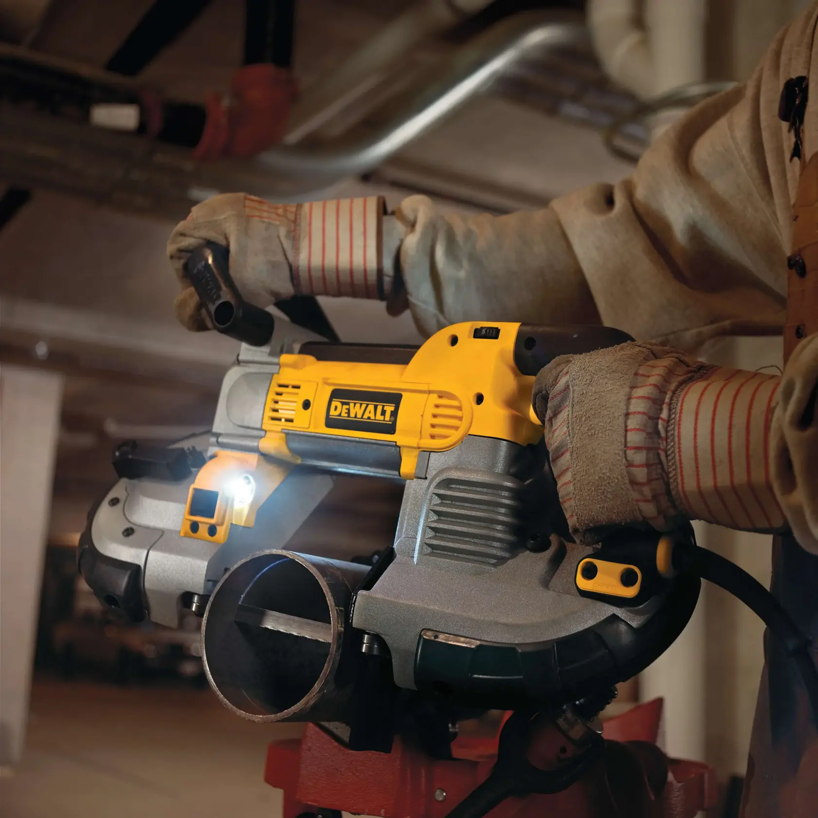 A person wearing protective gloves operates a DEWALT DWM120 portable band saw, cutting a metal pipe in an industrial setting. The tool features a built-in LED light for improved visibility.