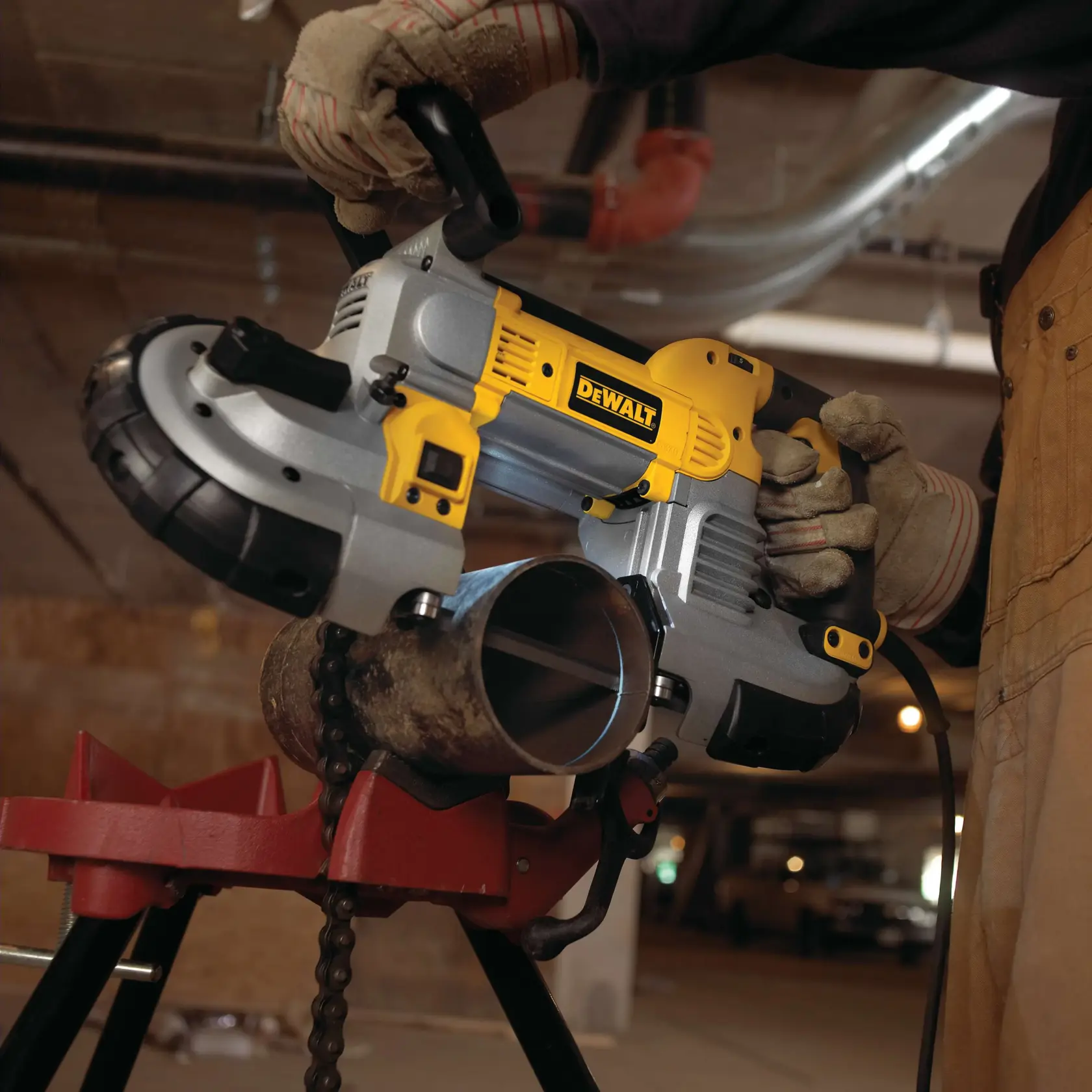 A person wearing gloves uses a DEWALT portable band saw to cut a metal pipe that is secured on a red stand in an industrial setting.