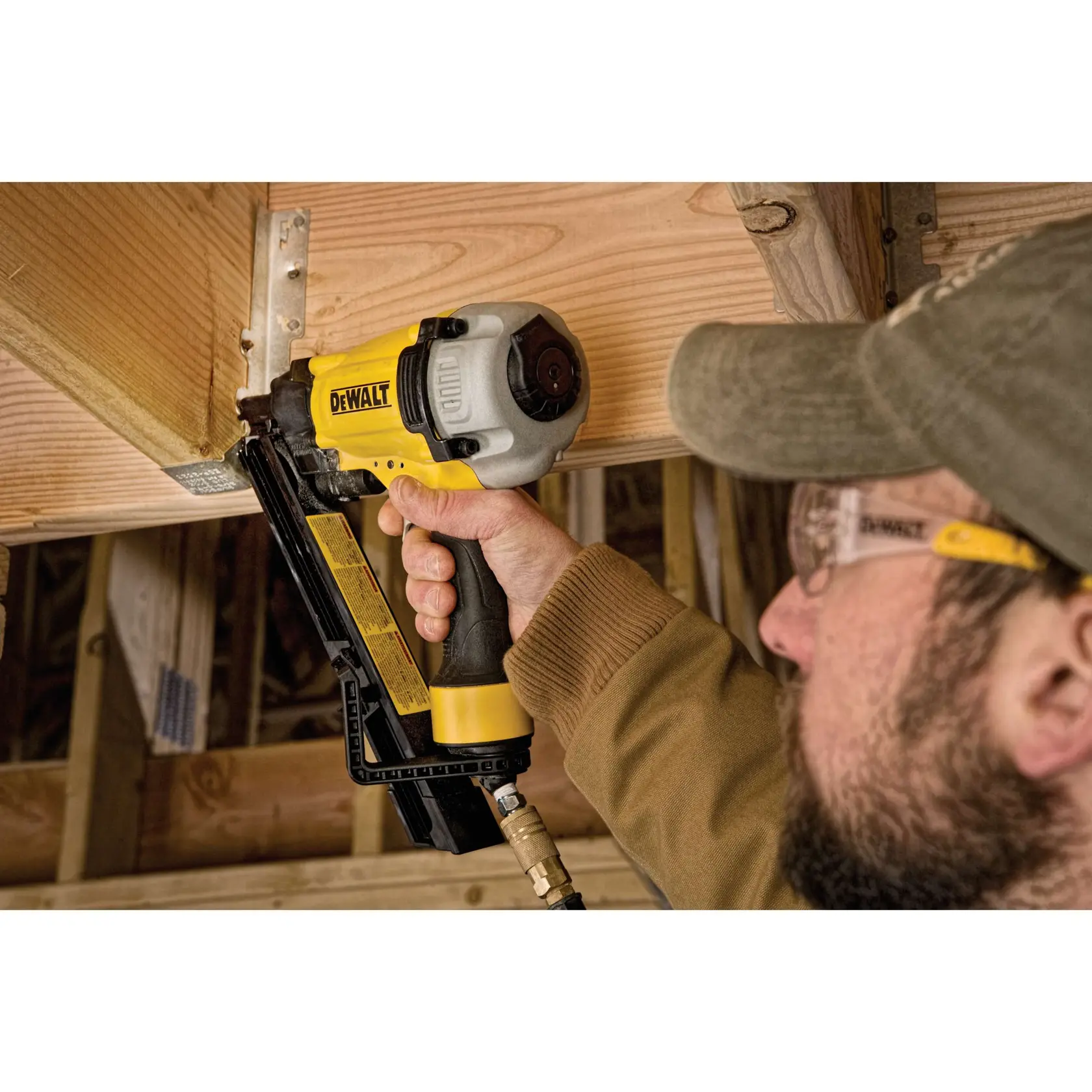 A person is using a DEWALT pneumatic nail gun to fasten wood beams during construction. The tool is yellow and black with a silver head. The person's face is blurred for privacy.