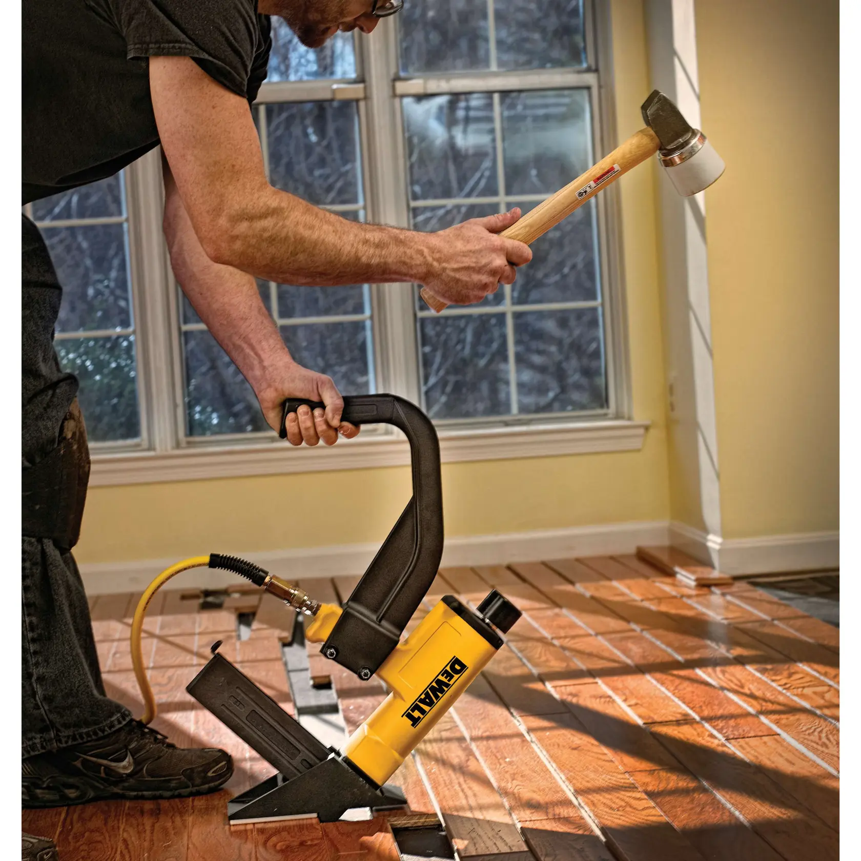 A person using a DEWALT pneumatic flooring nailer to install hardwood flooring in a room, holding a mallet and pressing down on the tool. Sunlight is coming through the windows in the background.