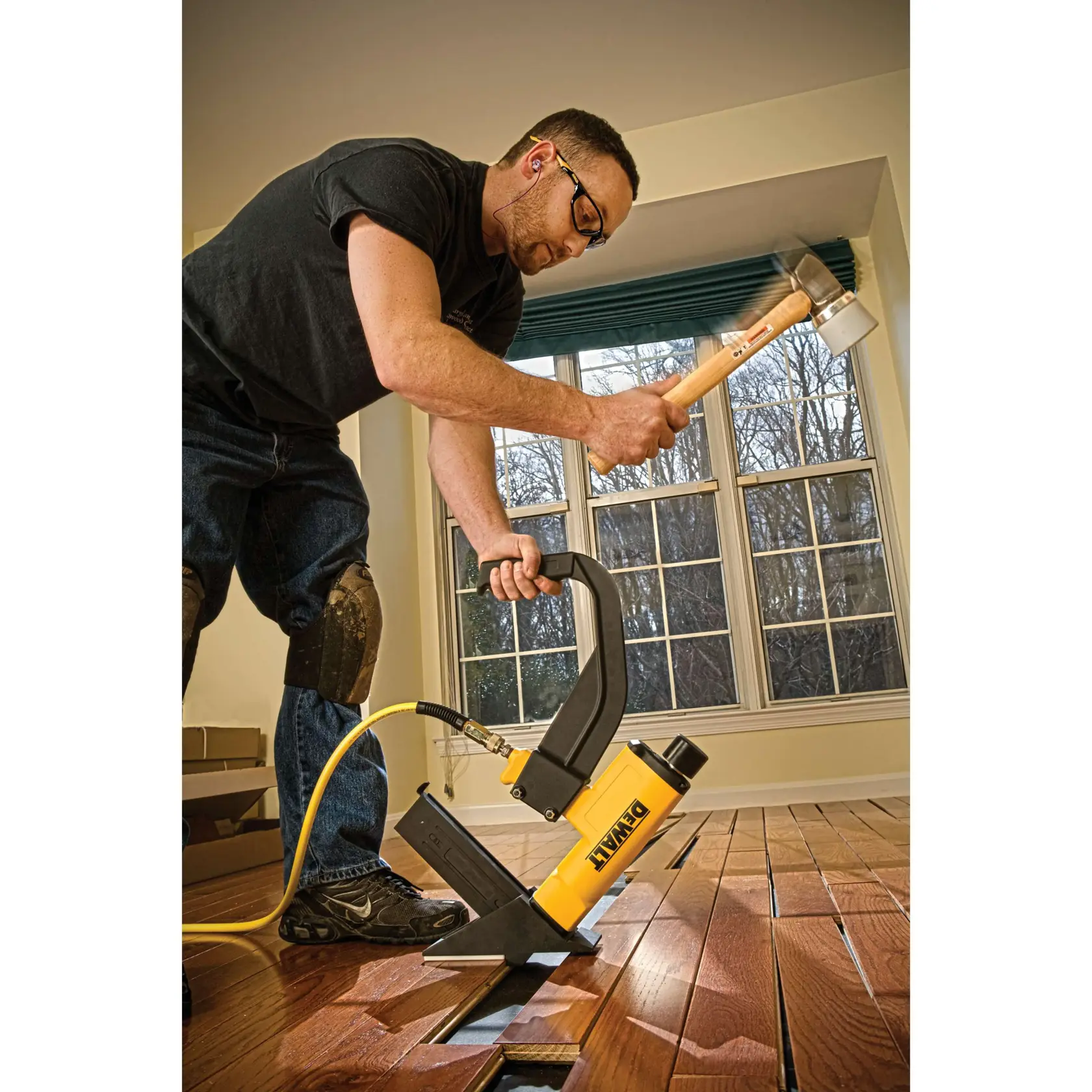 A person is installing hardwood flooring using a DEWALT flooring nailer and a mallet in a room with large windows. The person is wearing knee pads and using a yellow pneumatic nailer connected to an air hose.