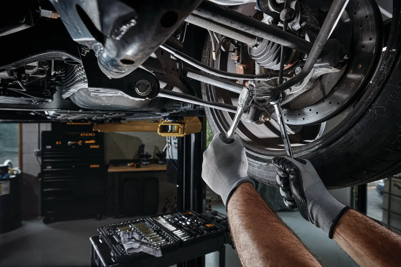 A person wearing work gloves uses wrenches to repair the suspension of a car, which is lifted on a hydraulic lift. A DEWALT mechanic tool set is visible in the foreground.