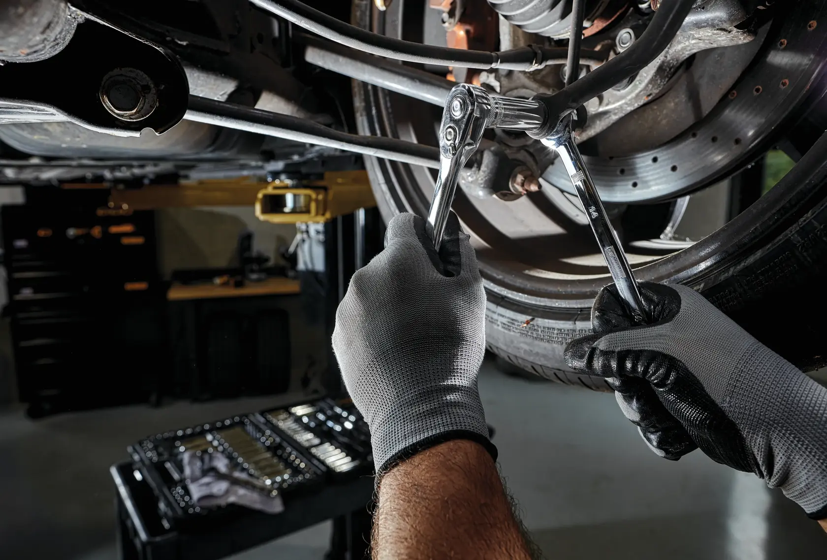 Close-up of gloved hands using DEWALT ratchets and wrenches to work on a vehicle's suspension or brake component, with a tool set visible in the background.