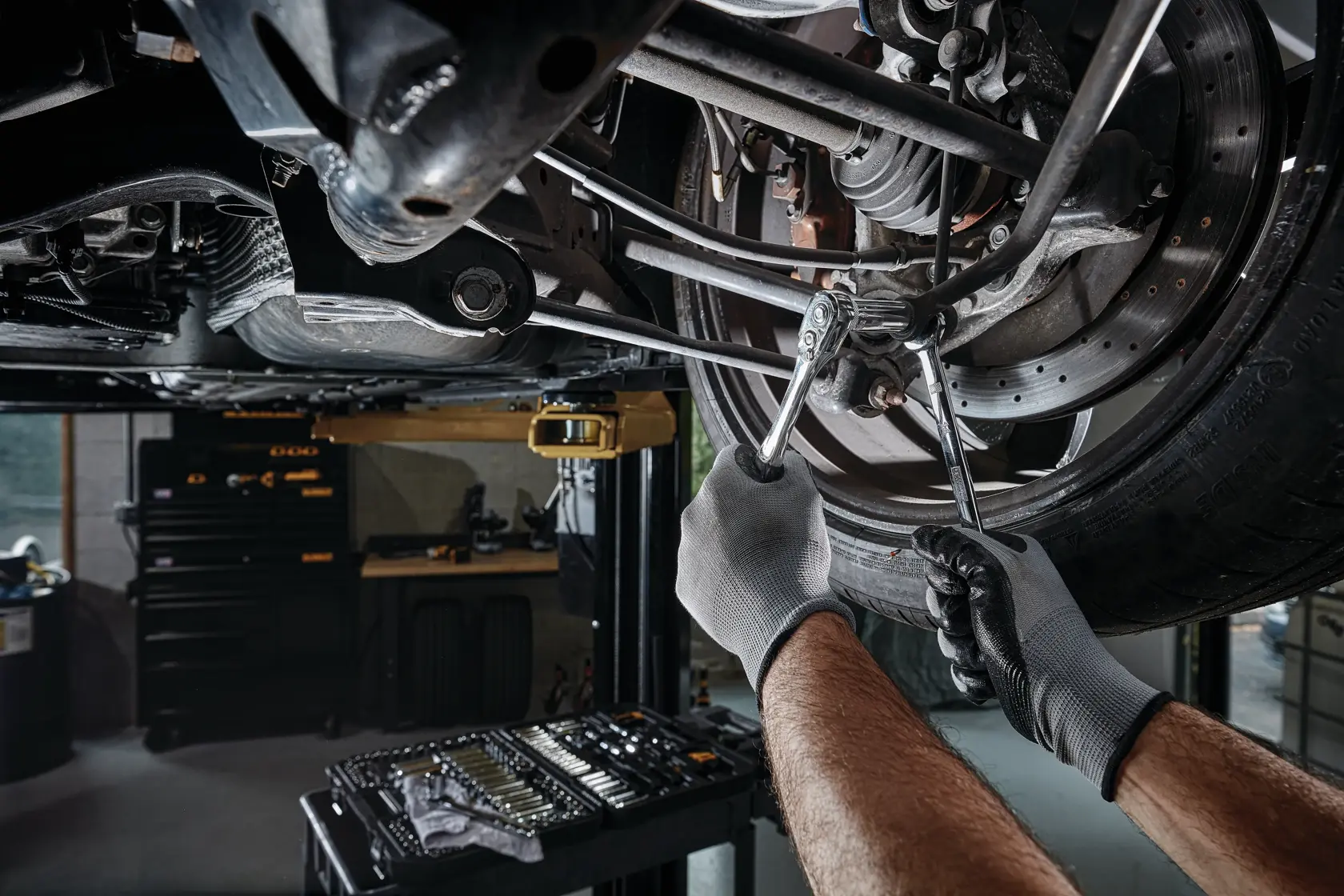 A person wearing gloves uses wrenches to perform maintenance on the suspension of a car elevated on a lift. A tool kit is visible on a table nearby in a garage setting.