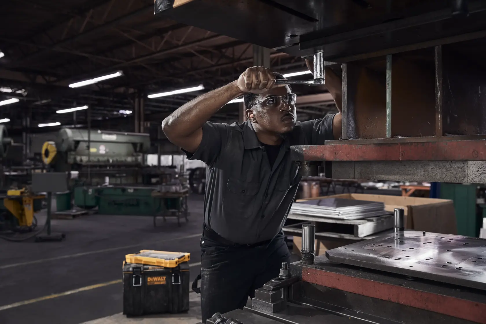 A person working in an industrial factory environment, operating machinery. In the foreground, there is a DEWALT toolbox on the floor. The factory workspace includes various equipment and metal materials.