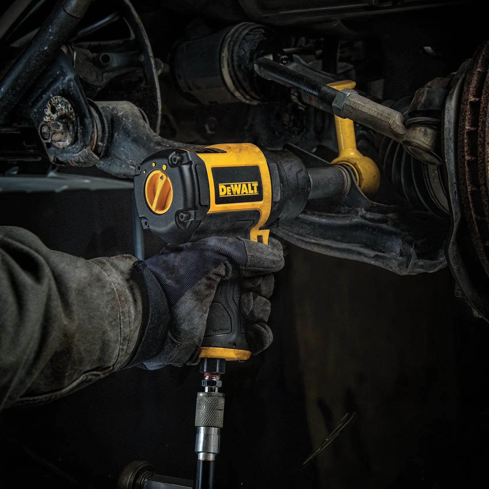 A person wearing work gloves is using a DEWALT impact wrench to work on a vehicle's suspension or axle component.