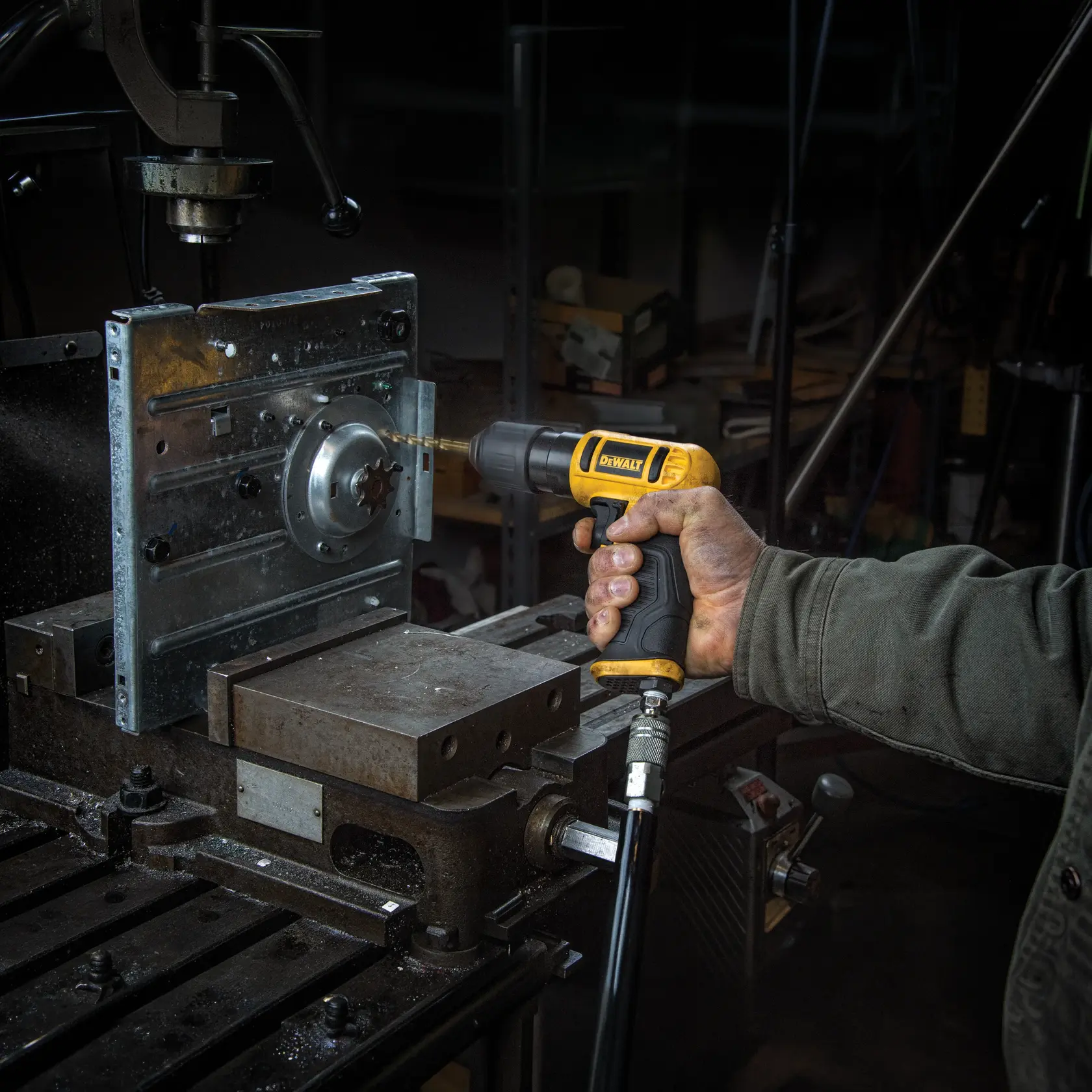 A person is using a DEWALT pneumatic drill to bore into a metal workpiece mounted in a workshop setting. The drill is connected to an air hose and the workspace features industrial equipment.