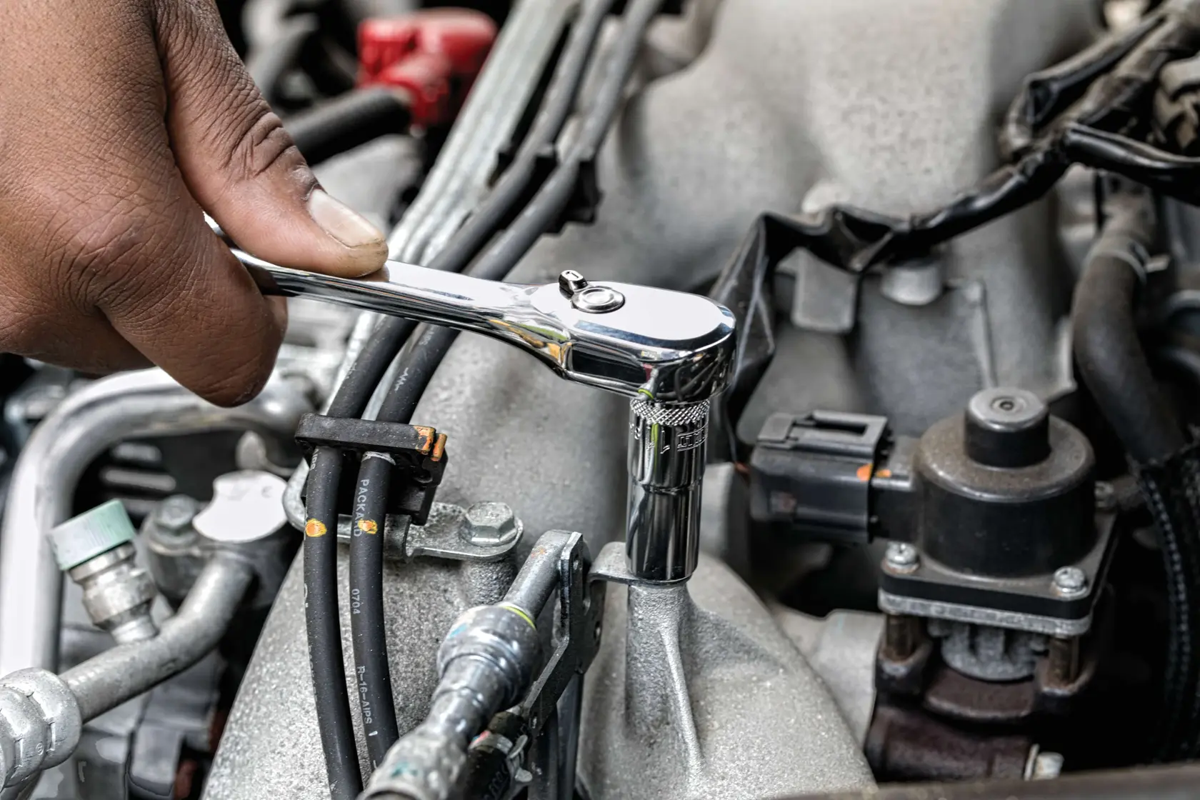 A hand is using a DEWALT ratchet wrench to tighten or loosen a bolt on an engine component, surrounded by various hoses and mechanical parts.