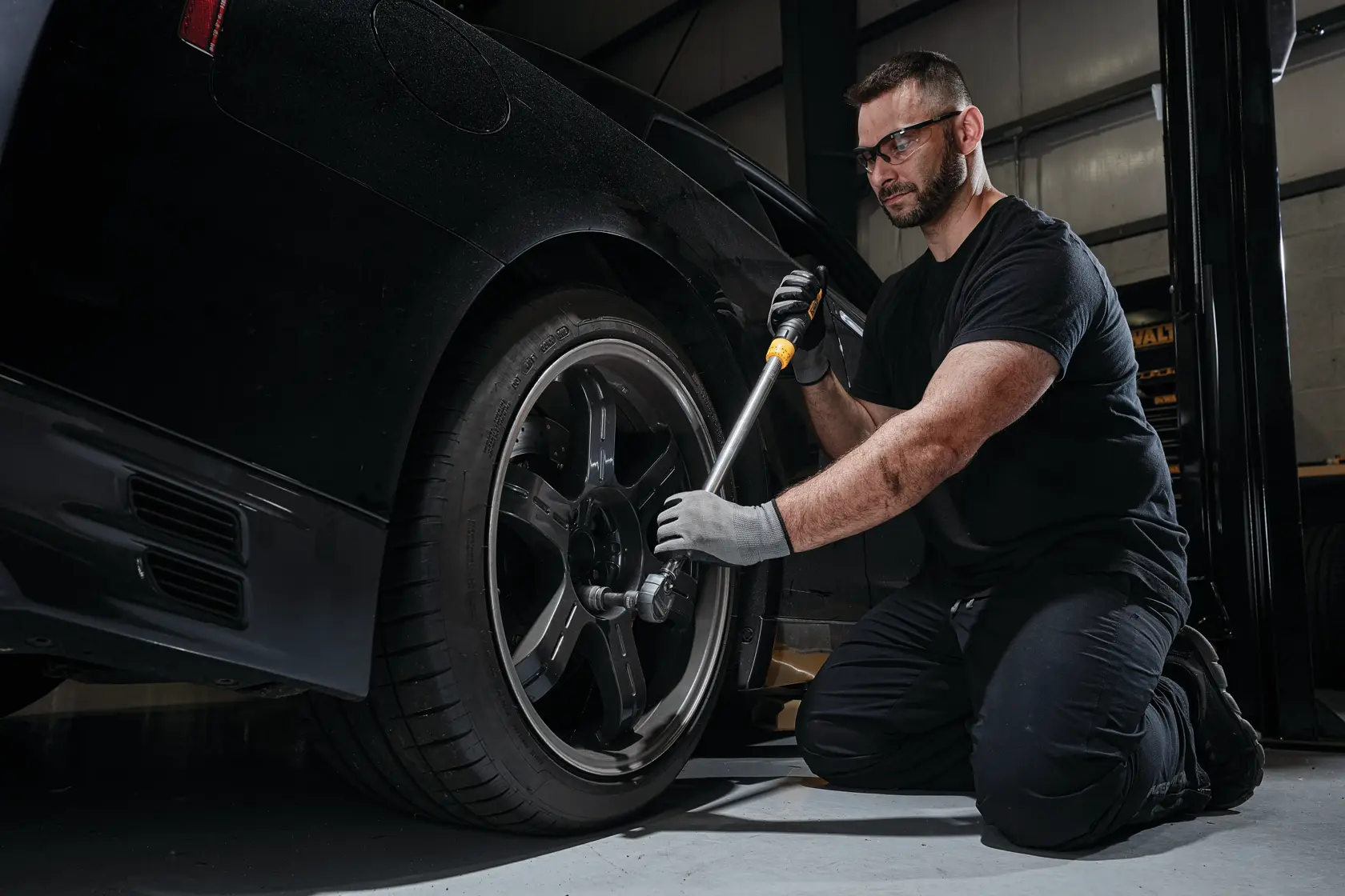 A person wearing gloves uses a DEWALT torque wrench to tighten the lug nuts on a car wheel in a garage. The individual is kneeling on the ground beside the vehicle.