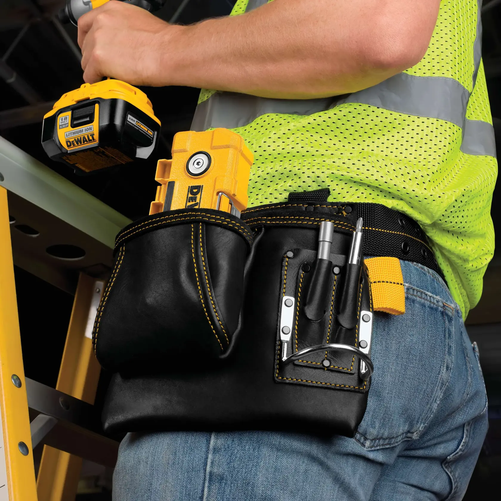 A close-up view of a person wearing a black leather tool belt with yellow stitching, containing various DEWALT tools and accessories. The person is also holding a DEWALT cordless power tool and is dressed in blue jeans and a bright yellow safety vest. A yellow ladder is visible in the background.