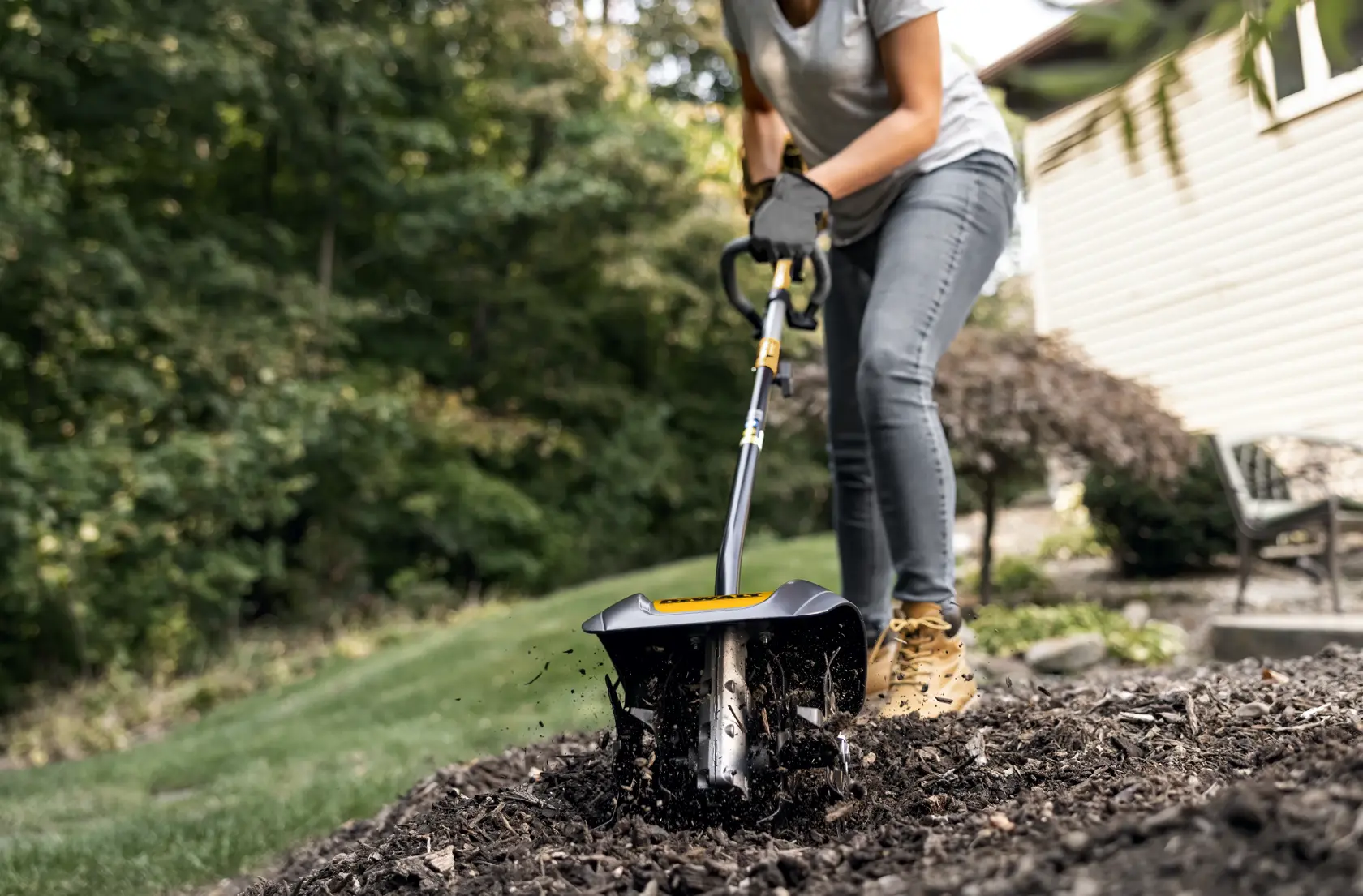 A person using a DEWALT garden cultivator attachment to till soil in a garden bed, with soil being turned up by the rotating blades.