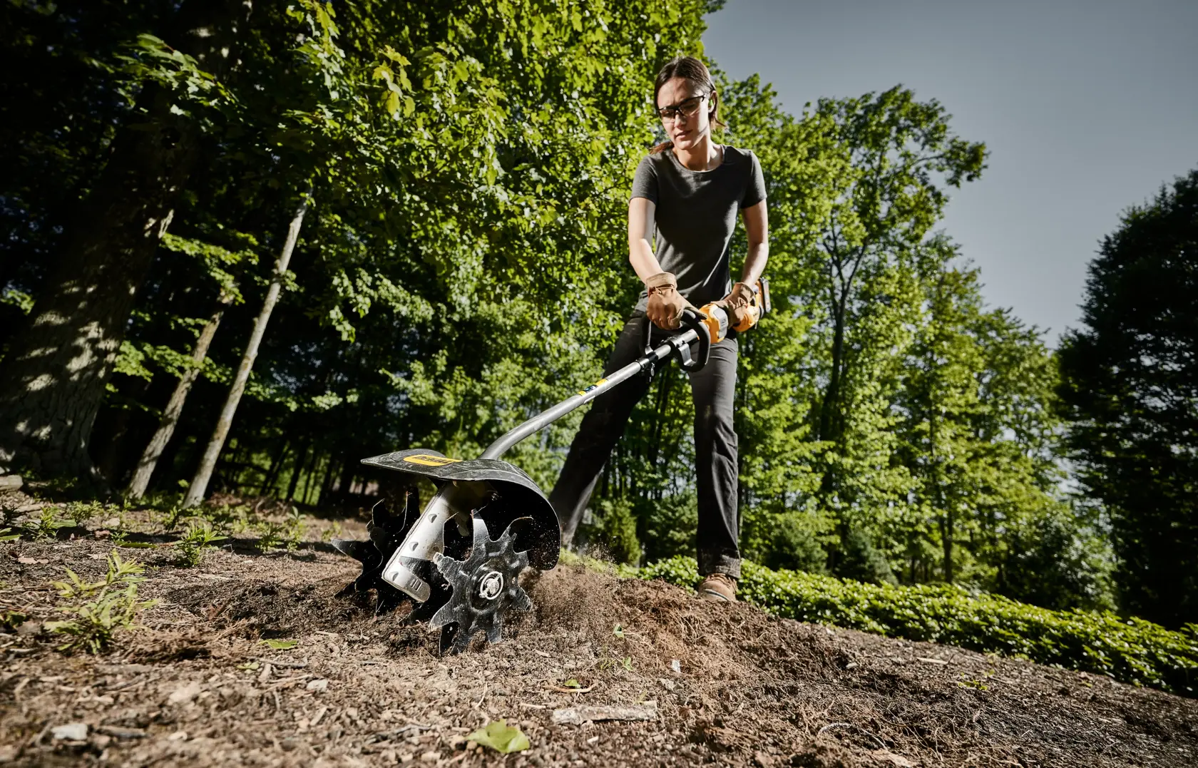 A person with a blurred face uses a DEWALT DWOAS3CL cultivator attachment outdoors, preparing soil in a garden area surrounded by trees.