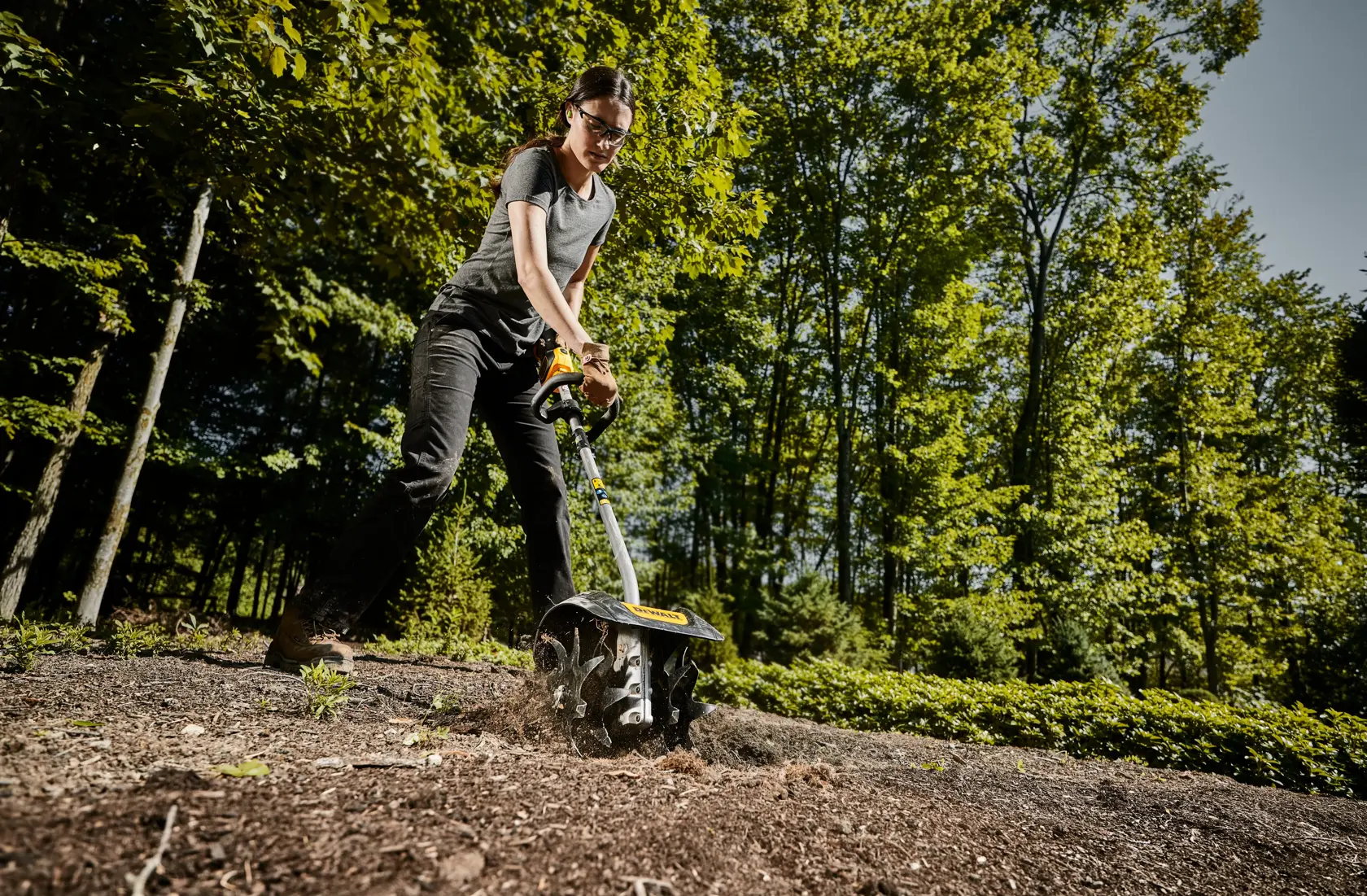 Person using a DEWALT garden cultivator attachment outdoors on soil, surrounded by green trees. The individual's face is blurred for privacy.