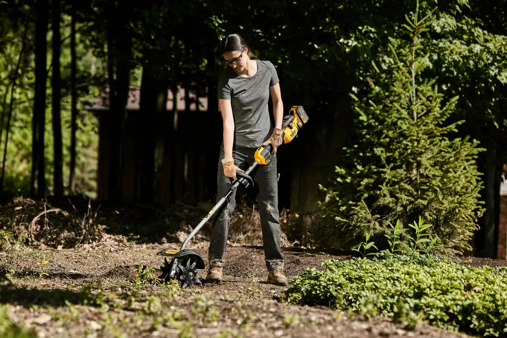 A person using a DEWALT cordless outdoor tool to cultivate soil in a garden area, wearing gloves and outdoor clothing with their face blurred for privacy. There are trees and greenery in the background.