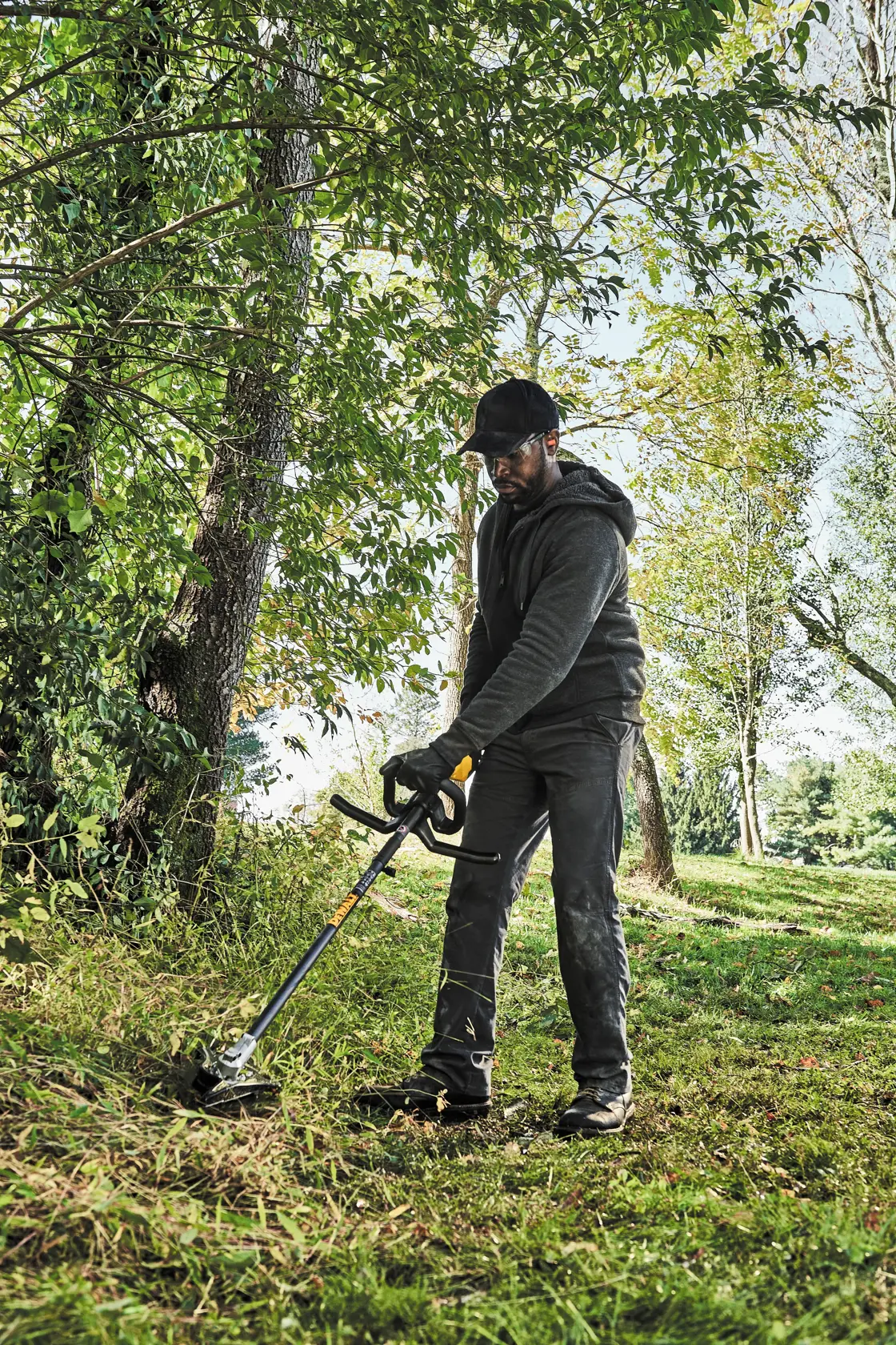 A person wearing a dark hoodie, pants, gloves, and a cap is using a DEWALT brush cutter attachment to trim grass and vegetation outdoors in a wooded area.