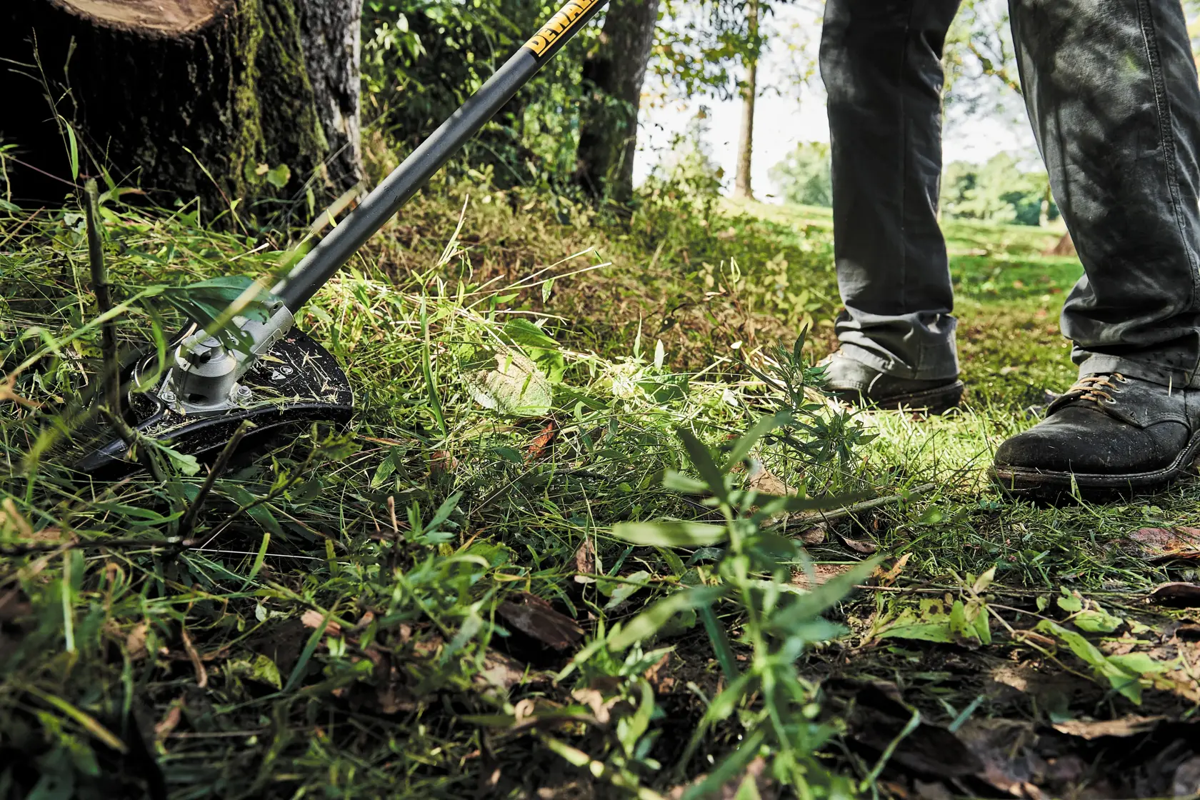 A person wearing work boots is using a DEWALT brush cutter attachment to trim grass and weeds in an outdoor area.