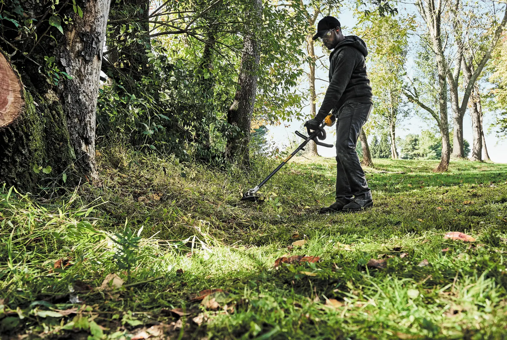 A person is using a DEWALT brush cutter to trim grass and weeds in a wooded outdoor area. The person is wearing work clothes and gloves, operating the tool near trees and overgrown vegetation.