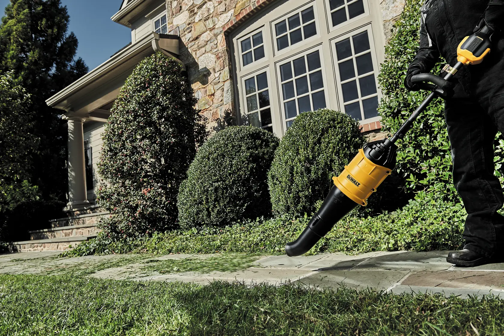 A person is using a DEWALT handheld leaf blower to clear grass clippings from a stone pathway in front of a house surrounded by trimmed bushes and greenery.