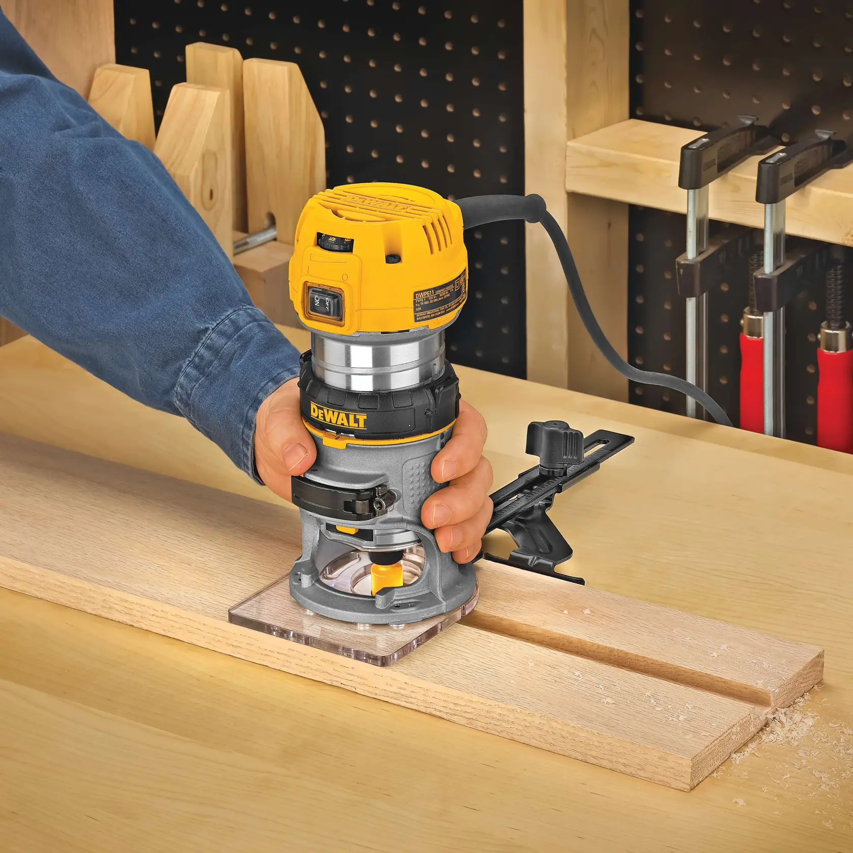 A person uses a DEWALT DWP611 wood router to carve grooves into a wooden board on a workbench. The router is powered and clamped for precision. Workshop tools and clamps are visible in the background.
