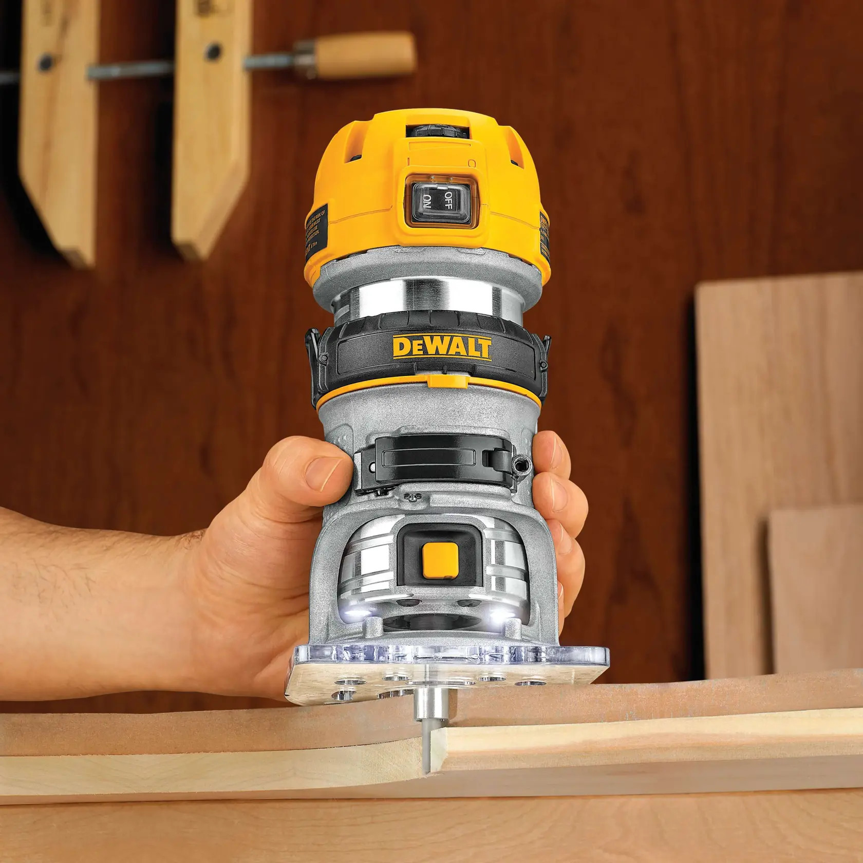 A hand holding a DEWALT DWP611PK compact router, working on a wooden edge. The router is yellow and silver with a visible power button and LED lights at the base. Wood clamps and wooden boards are in the background.