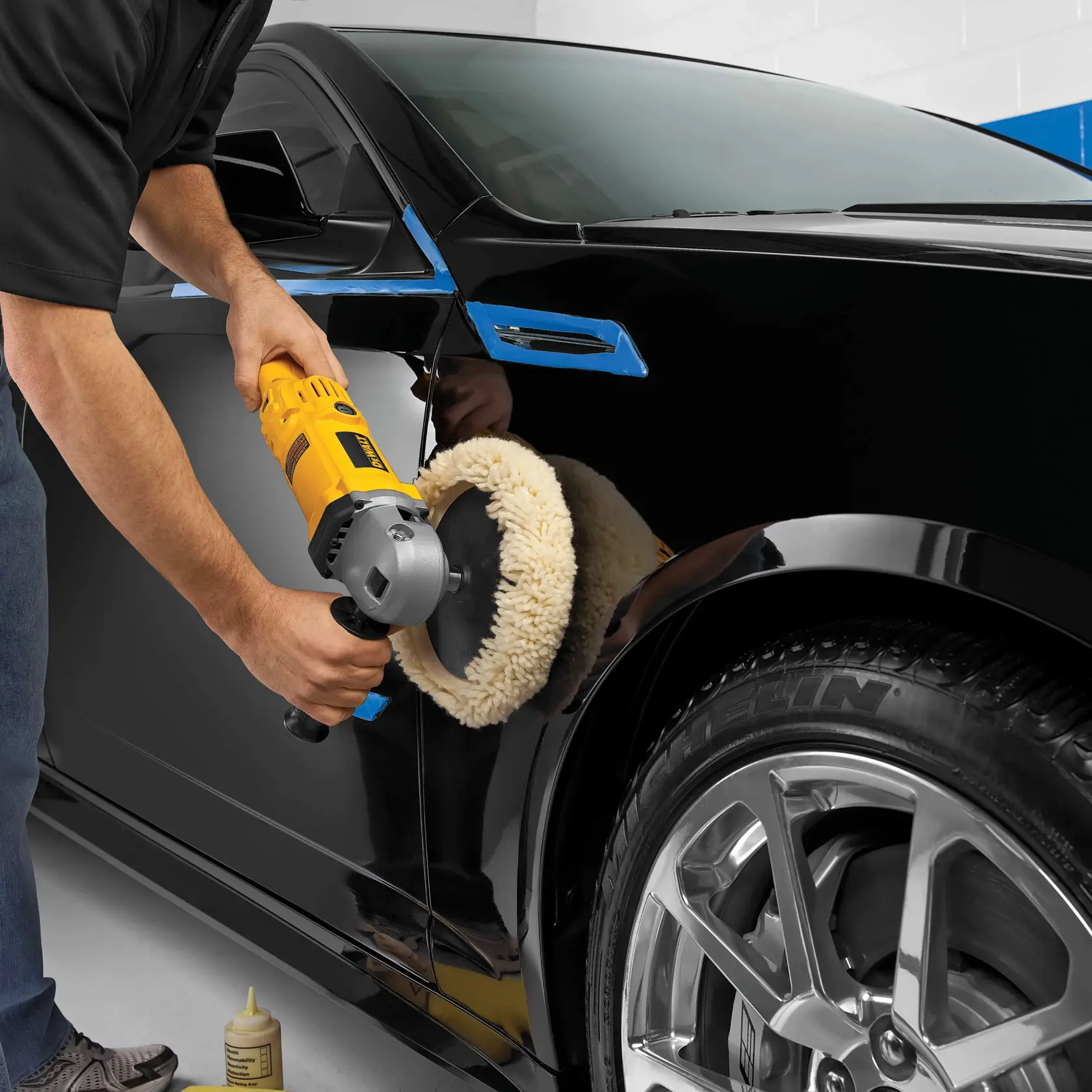 A person is using a DEWALT DWP849 rotary polisher with a wool pad to polish the exterior of a black car. The car's surface is shiny, and the polisher is being held firmly against the paintwork. Polishing solution is visible on the floor nearby.