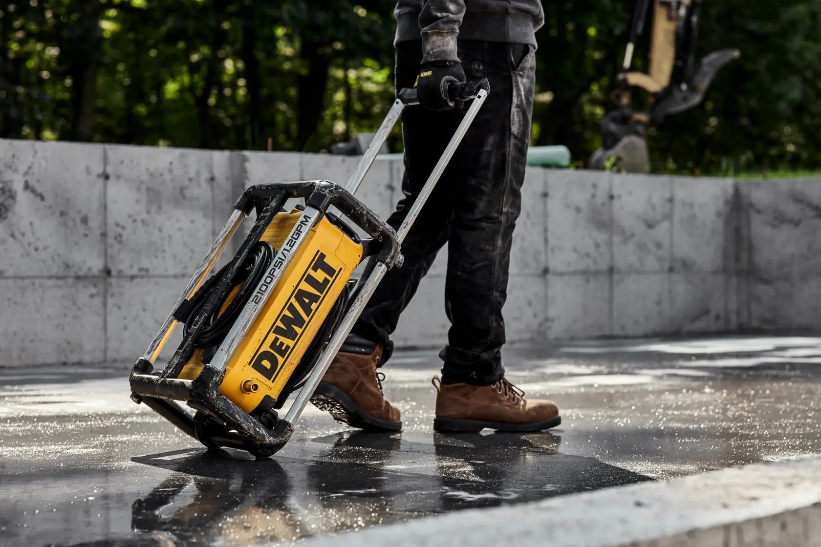 A person wearing work boots is pulling a yellow DEWALT portable power washer with wheels and a handle on a wet concrete surface outdoors.