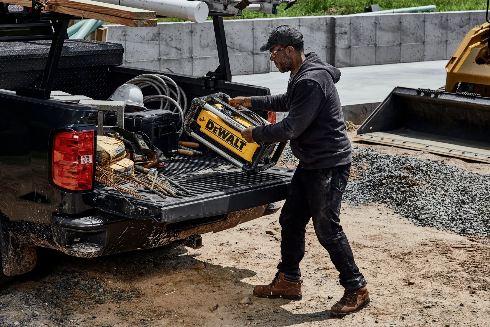 A person is loading a DEWALT DWPW2100 power washer into the bed of a pickup truck at a construction site. The truck bed contains various tools and construction materials.