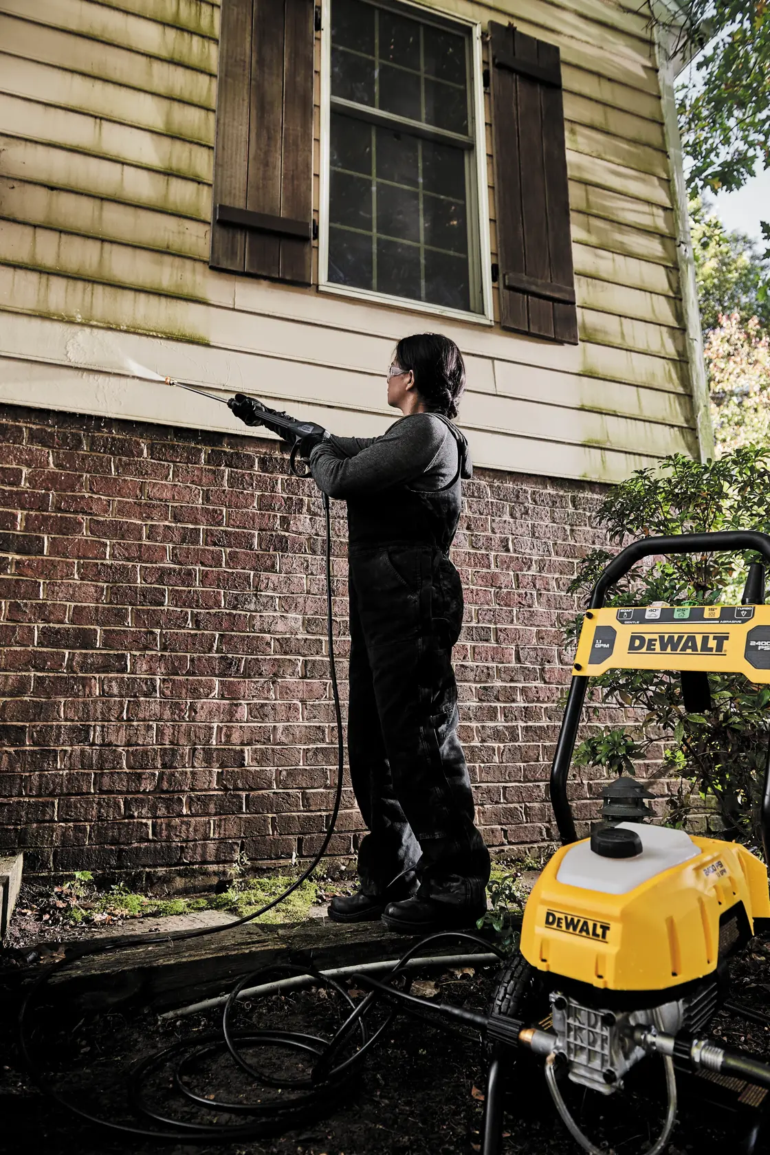 A person is using a DEWALT pressure washer to clean the siding of a house. The house has brick on the lower part and light-colored siding above, with some visible green stains being cleaned off. The pressure washer is yellow and branded with the DEWALT logo.