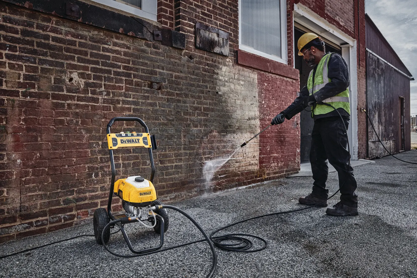 A person wearing a safety vest and helmet uses a DEWALT pressure washer to clean the exterior brick wall of a building.