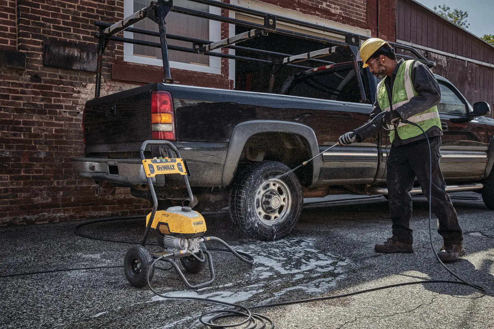 A person wearing safety gear uses a DEWALT DWPW2400 pressure washer to clean the wheel of a pickup truck parked outdoors. The pressure washer is positioned on the pavement with soap and water visible around the truck.