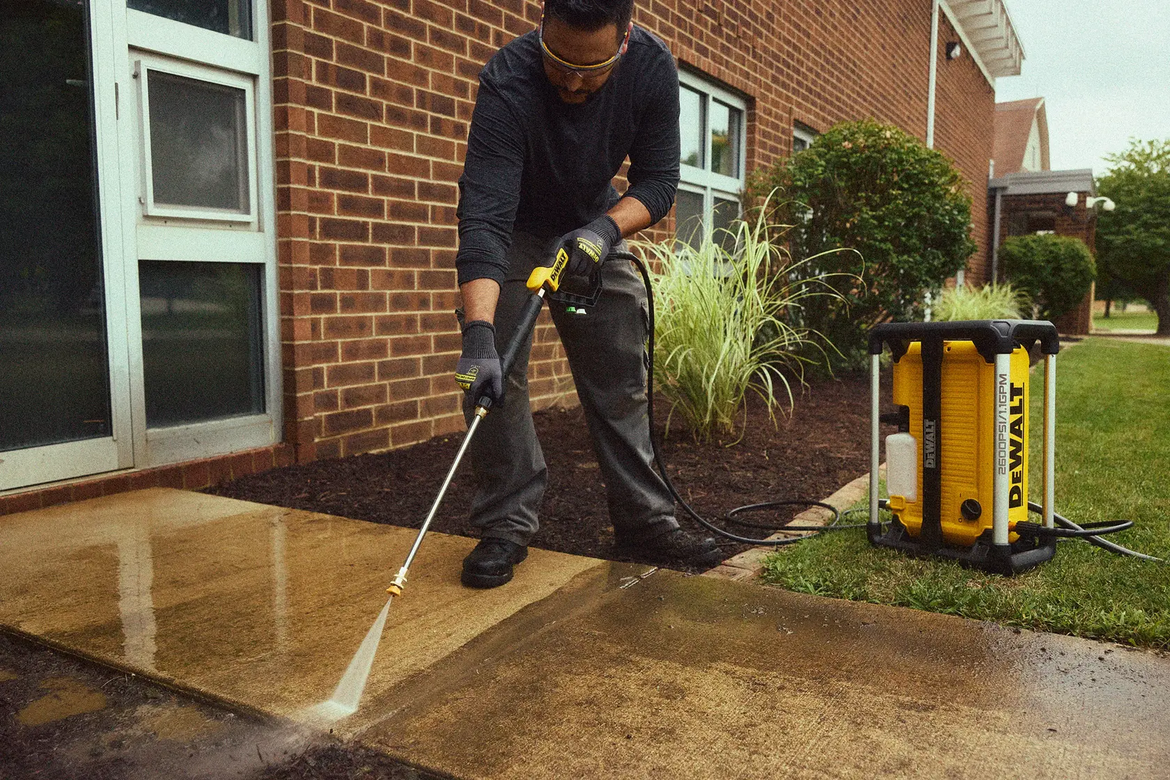A person is using a DEWALT DWPW2600 pressure washer to clean a concrete sidewalk outside a brick building. The pressure washer is positioned on the grass beside the walkway, connected with a hose, and water is being sprayed to remove dirt.
