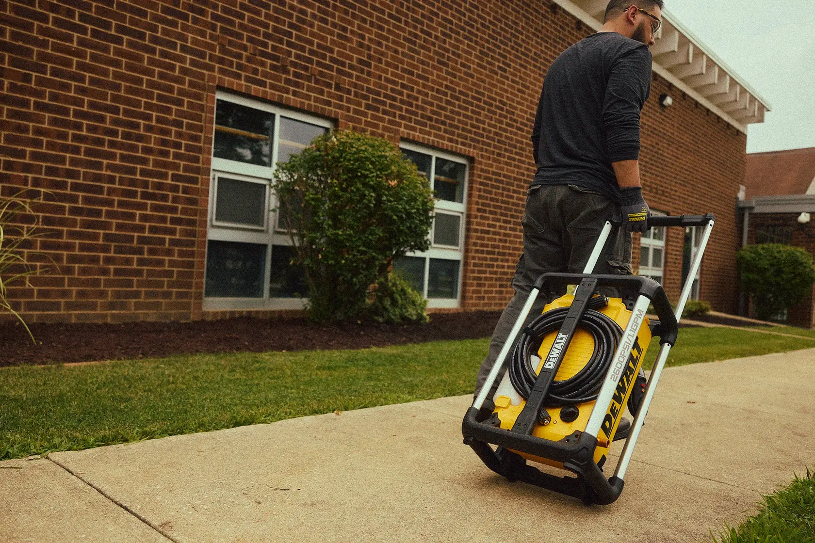 A person is pulling a DEWALT portable pressure washer with wheels and a handle along a sidewalk outside a brick building.