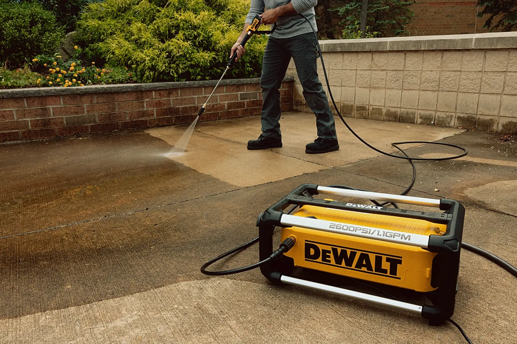 A person is using a DEWALT DWPW2600 pressure washer to clean an outdoor concrete surface, with the yellow pressure washer prominently placed in the foreground.